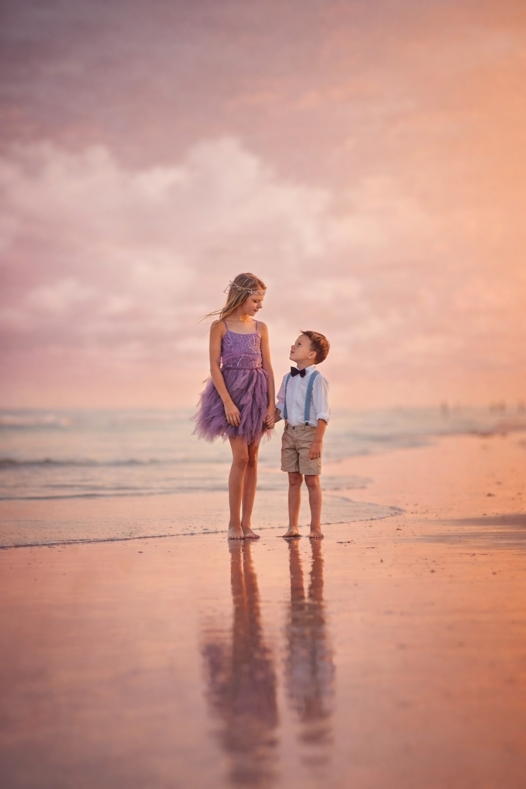 An older sister and younger brother stand together on reflective wet sand as the ocean meets the horizon at sunset.