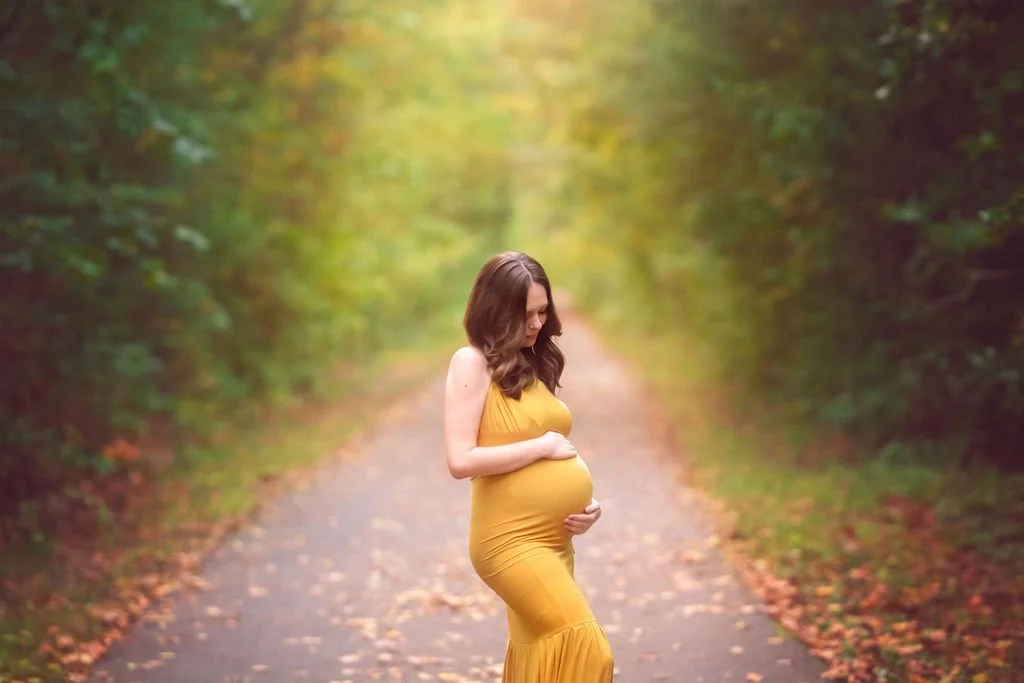 Pregnant mother in a yellow dress looking down at her baby bump during an outdoor maternity photography session in South Jersey.