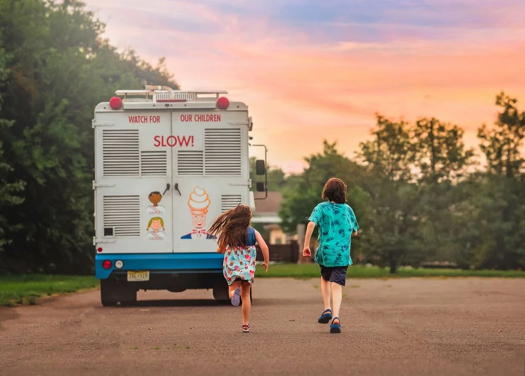 Children enjoying their ice cream cones and running towards the Mister Softee truck in the background.