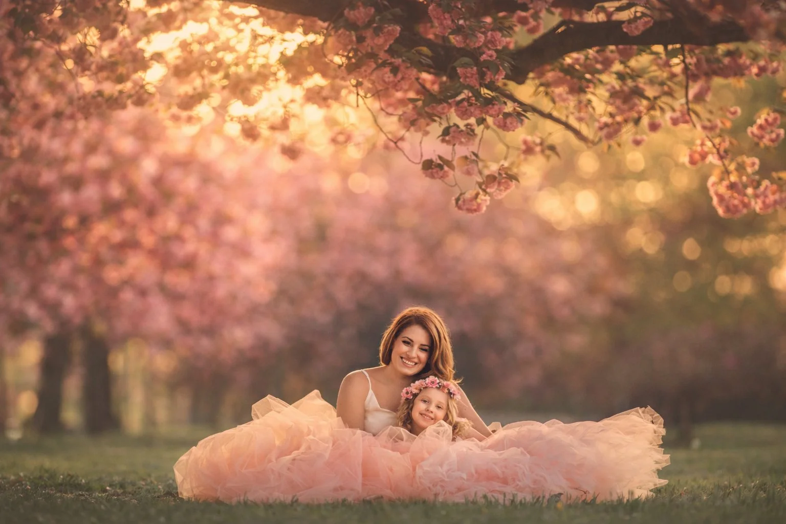 Mother and little girl sitting in the grass beneath cherry blossom trees and smiling during a spring portrait session in New Jersey.