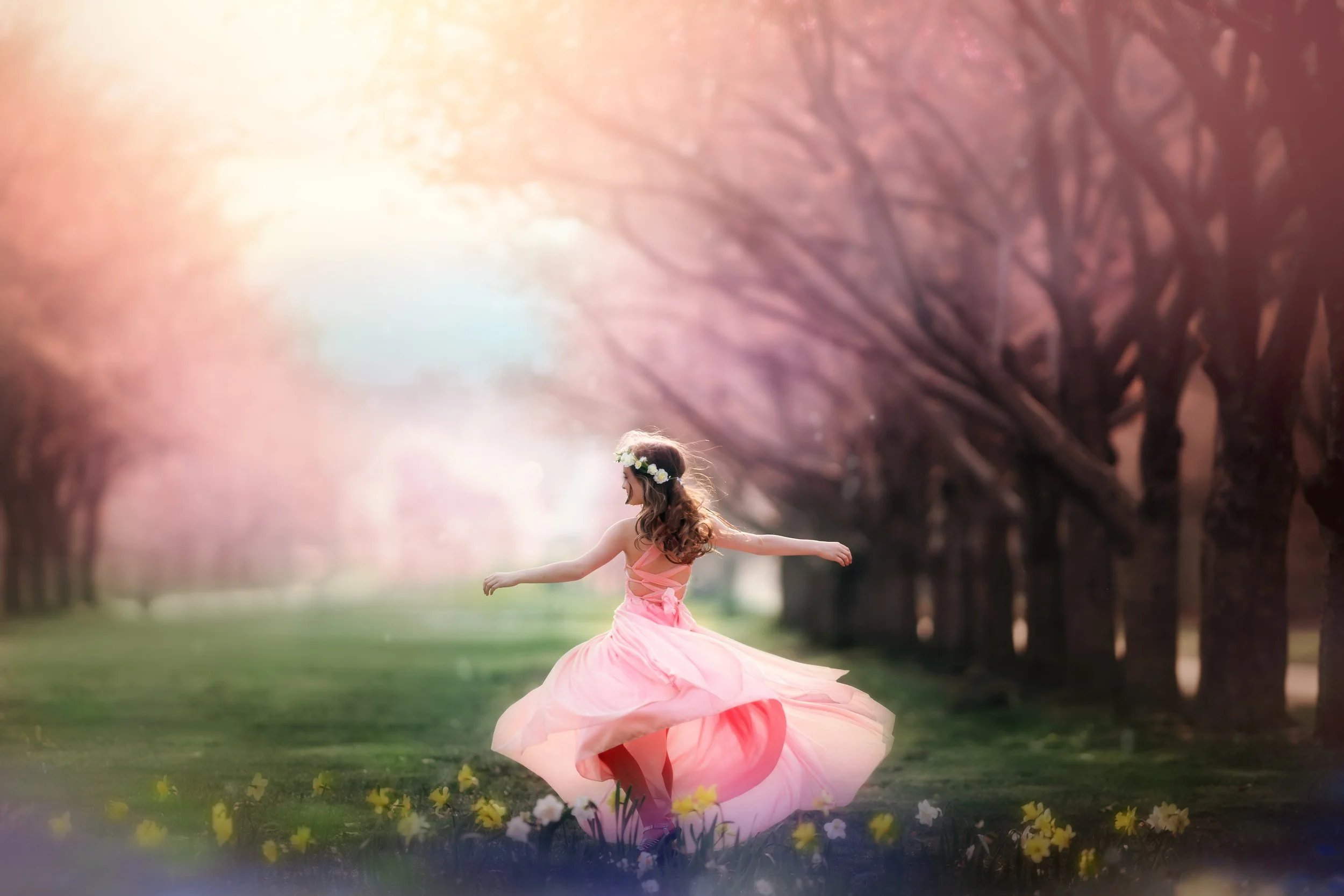 A young girl in a flowing pink dress twirls under soft cherry blossom trees during a fine art portrait session in South Jersey