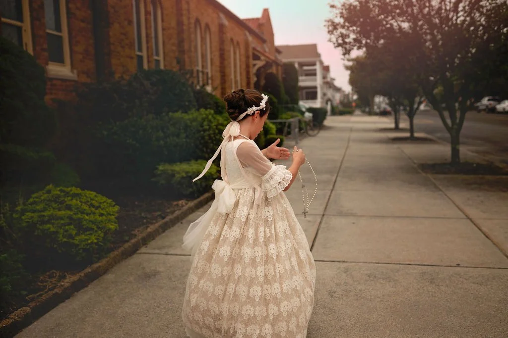 First Holy Communion portrait of a young girl photographed outside her church, standing on the sidewalk beside the church building