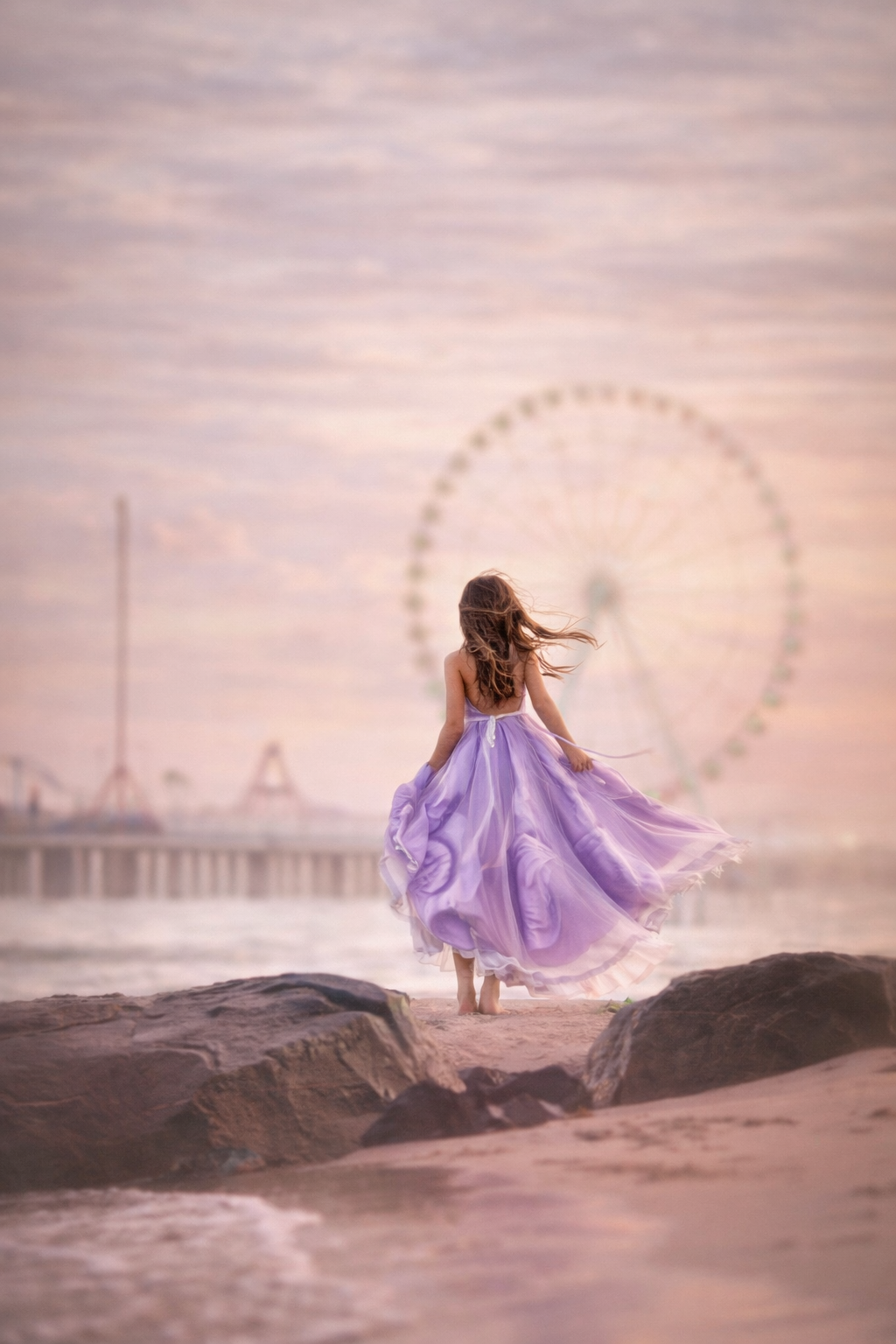 A girl in a flowing purple dress stands on seaside rocks with her back to the camera as the Ferris wheel fades into the distance.