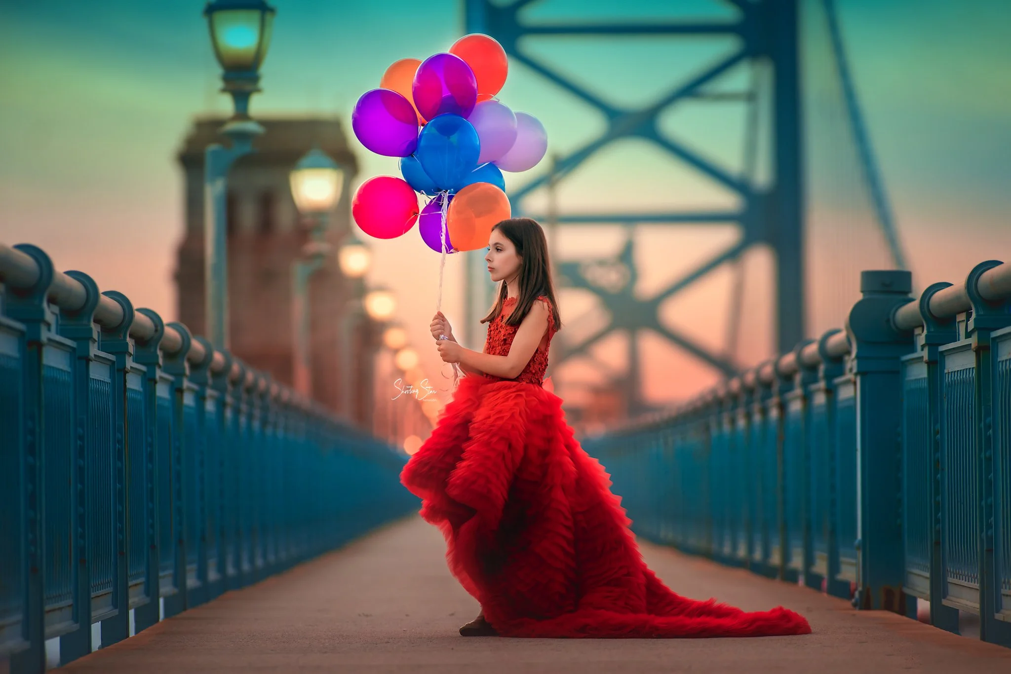 A fine art portrait of a young girl in a flowing red dress holding colorful balloons along a softly lit walkway. The image blends childhood wonder with editorial styling, creating a timeless, whimsical moment that feels cinematic and story-driven. 