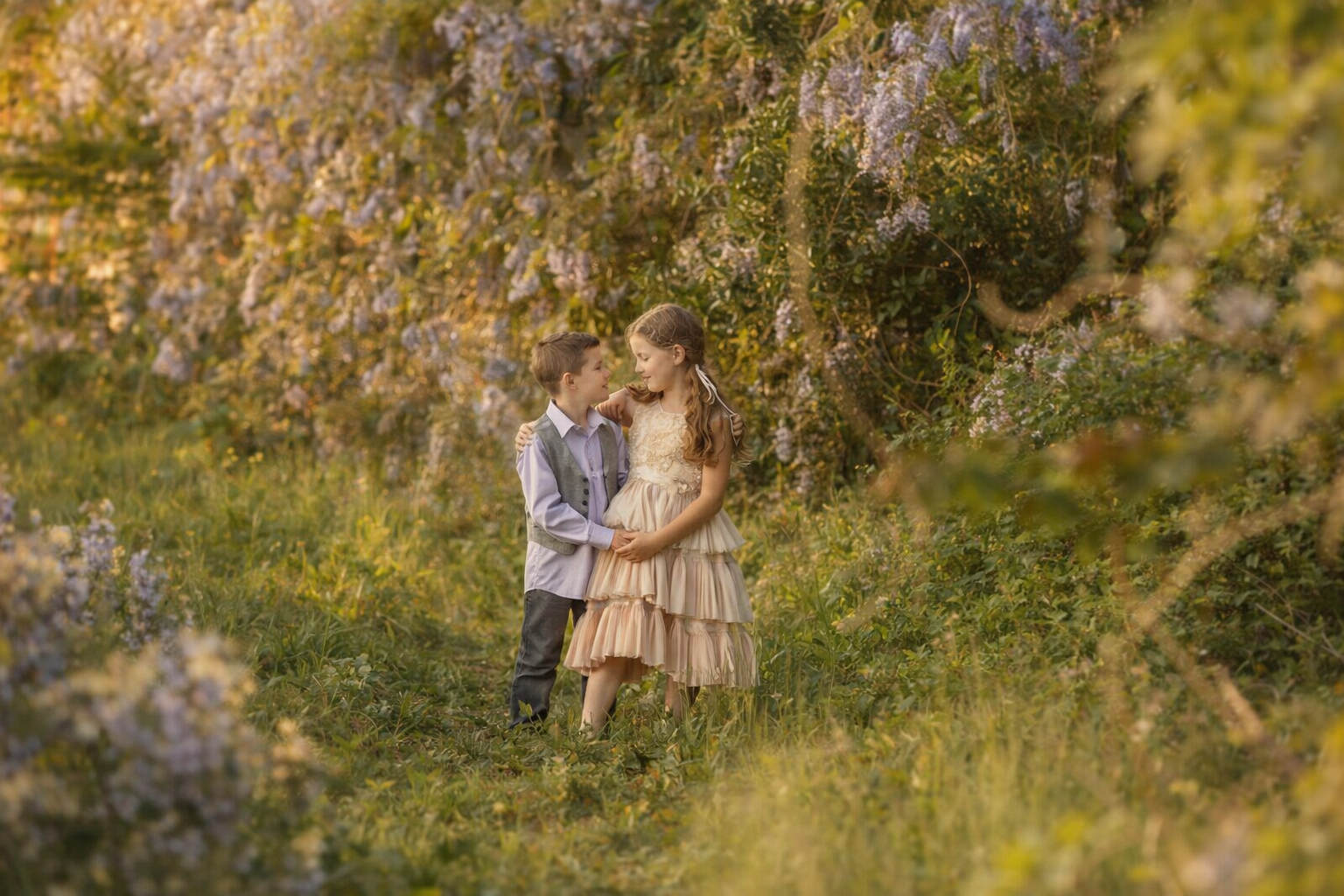 Brother and sister walking hand in hand under cascading purple wisteria during a timeless spring portrait session