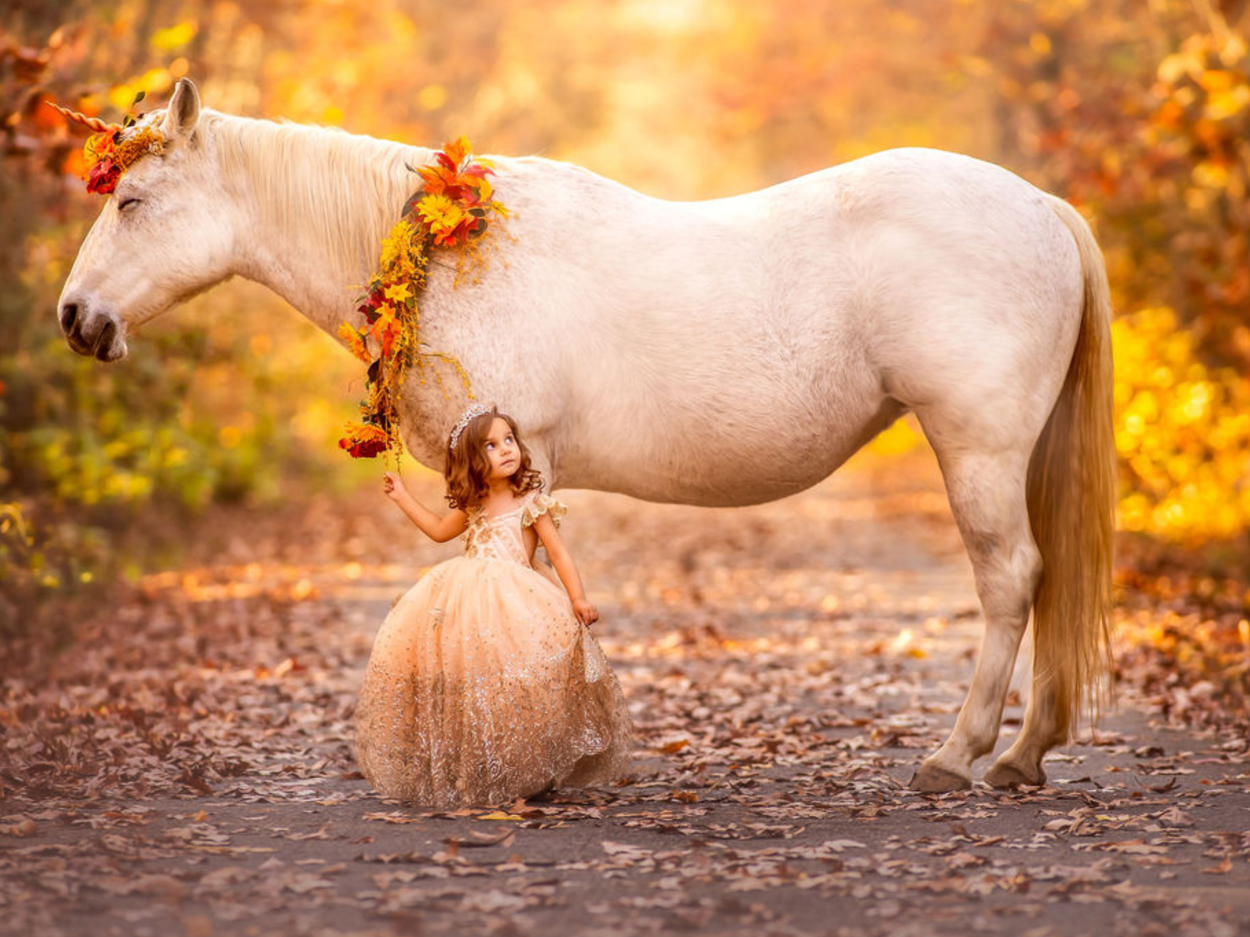 A young girl in a gold-toned dress stands beside a real unicorn on a leaf-covered forest path in fall, photographed as a fine art unicorn portrait experience in New Jersey