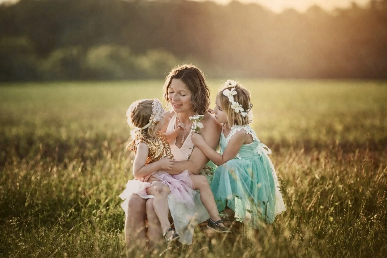 Mother sitting with her two young daughters in a sunlit field, fine art motherhood and family portrait
