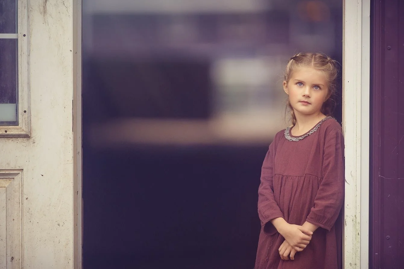 A young girl framed by an open barn door, surrounded by warm textures and natural light, creating a timeless fine art portrait inspired by rural simplicity and childhood wonder in South Jersey.