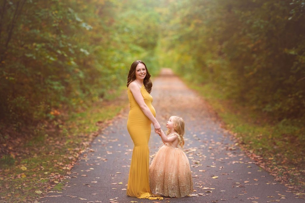 Pregnant mother smiling at the camera while her young daughter looks up at her during an outdoor maternity photography session in South Jersey.