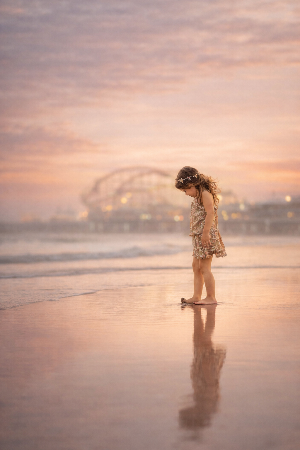 A quiet fine art portrait of a young girl standing alone on wet sand with the pier softly fading into the background.