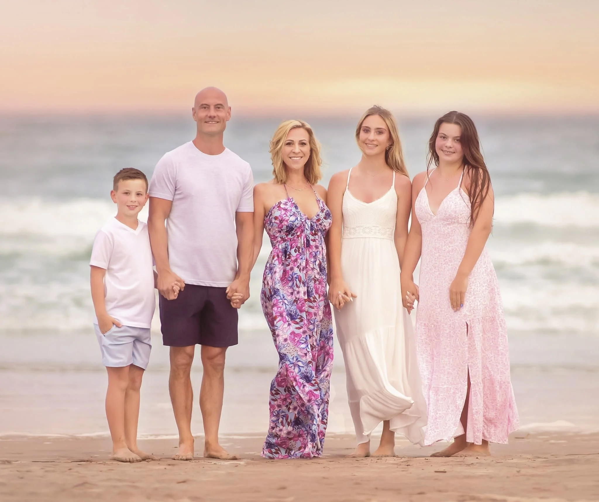 A family stands together facing the camera on the beach, framed by gentle waves and warm sunset light.