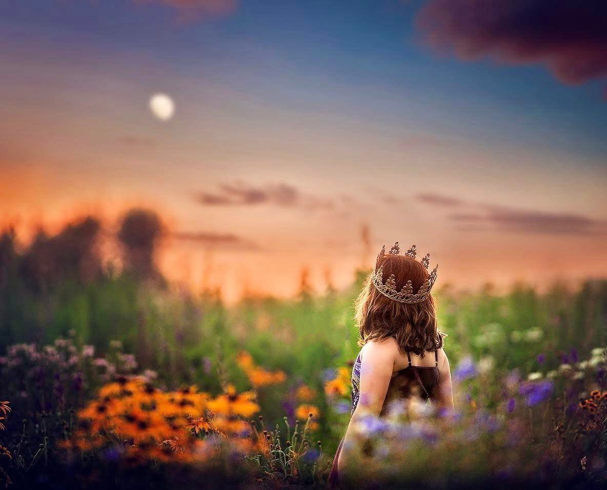 Girl wearing a crown standing in a wildflower field at sunset, looking toward the rising moon during a princess portrait session
