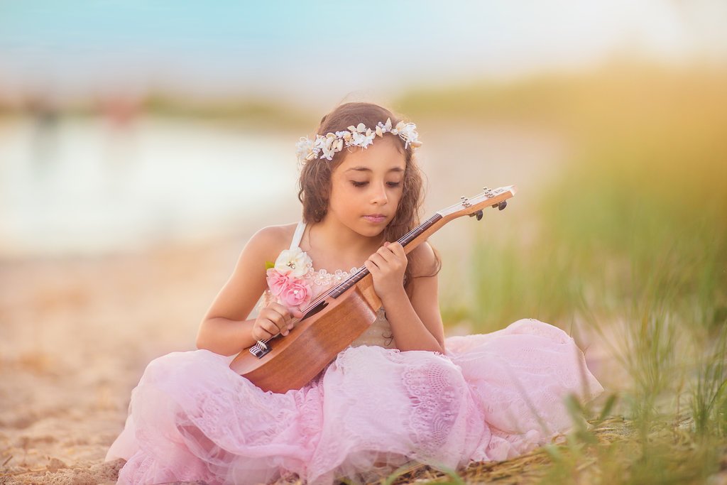 Little girl in a pink dress and seashell crown playing the guitar while sitting for portraits at the beach at the Jersey Shore, creating a whimsical and magical moment.