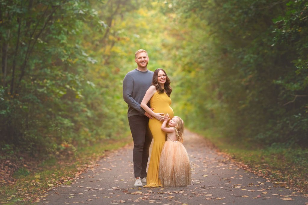 Family maternity photo with mom, dad, and young daughter smiling together outdoors during a South Jersey maternity session.