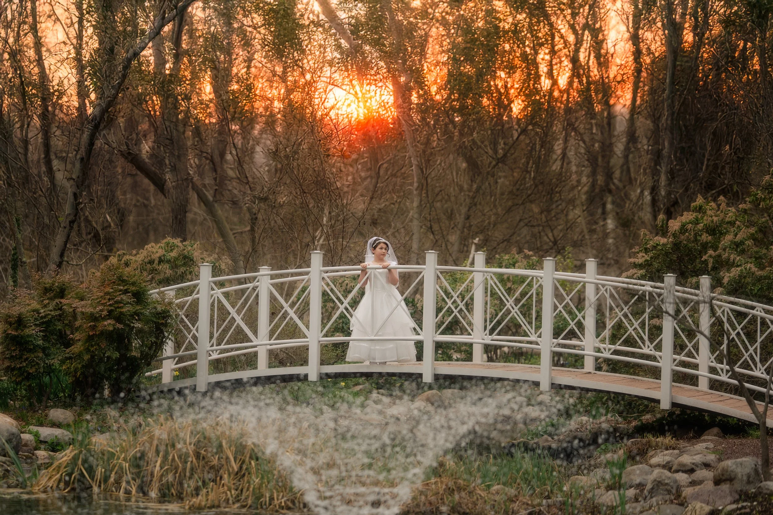 A girl in her first holy communion dress and veil standing on a small white bridge in a wooded area during sunset.