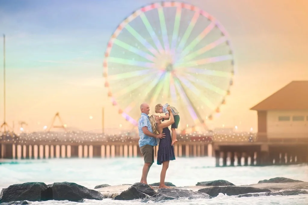 Mom lifting her little boy while the family poses on the beach in front of the Ferris wheel at Steel Pier in Atlantic City NJ.