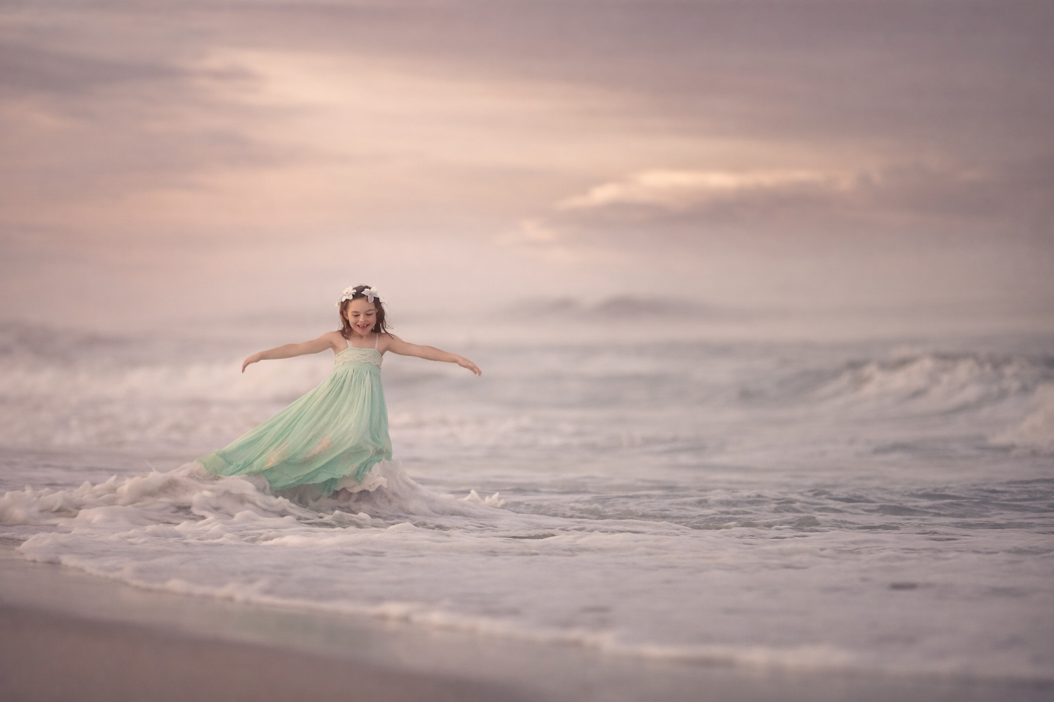 A young girl in a green dress photographed in the ocean in soft natural light for a timeless fine art children’s portrait.