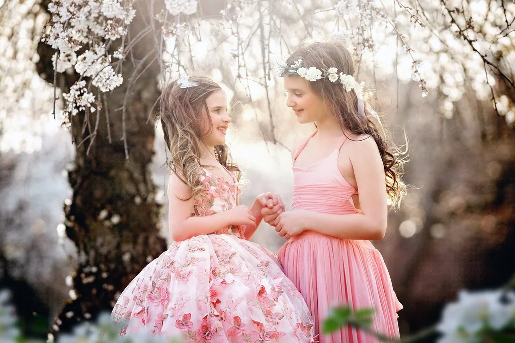 Two sisters looking at each other under soft spring blossoms during a children’s portrait session in Philadelphia