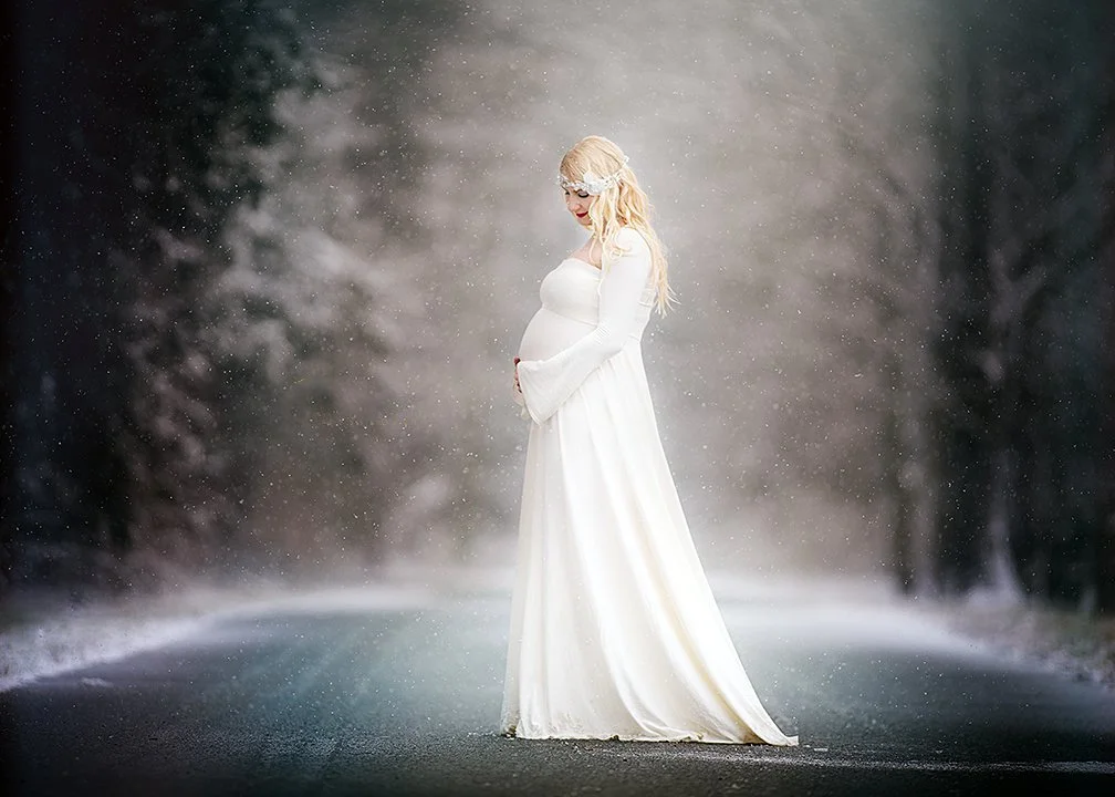 A pregnant woman in a white dress standing on an empty snowy road in a winter forest, her hands resting on her belly.