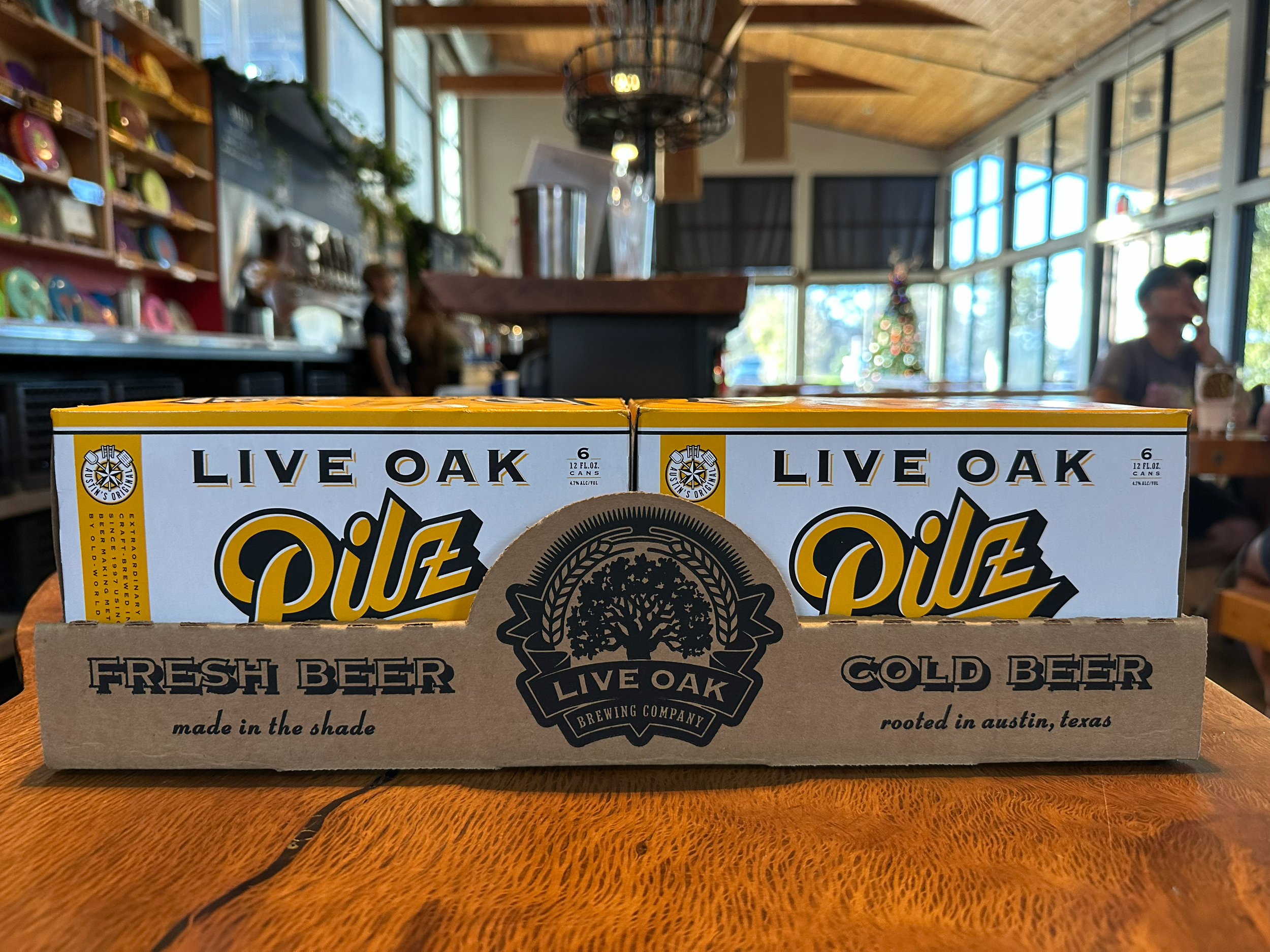 Person dressed in gray jeans and a black jacket walking outdoors while carrying a box of Live Oak Kalt IPA.