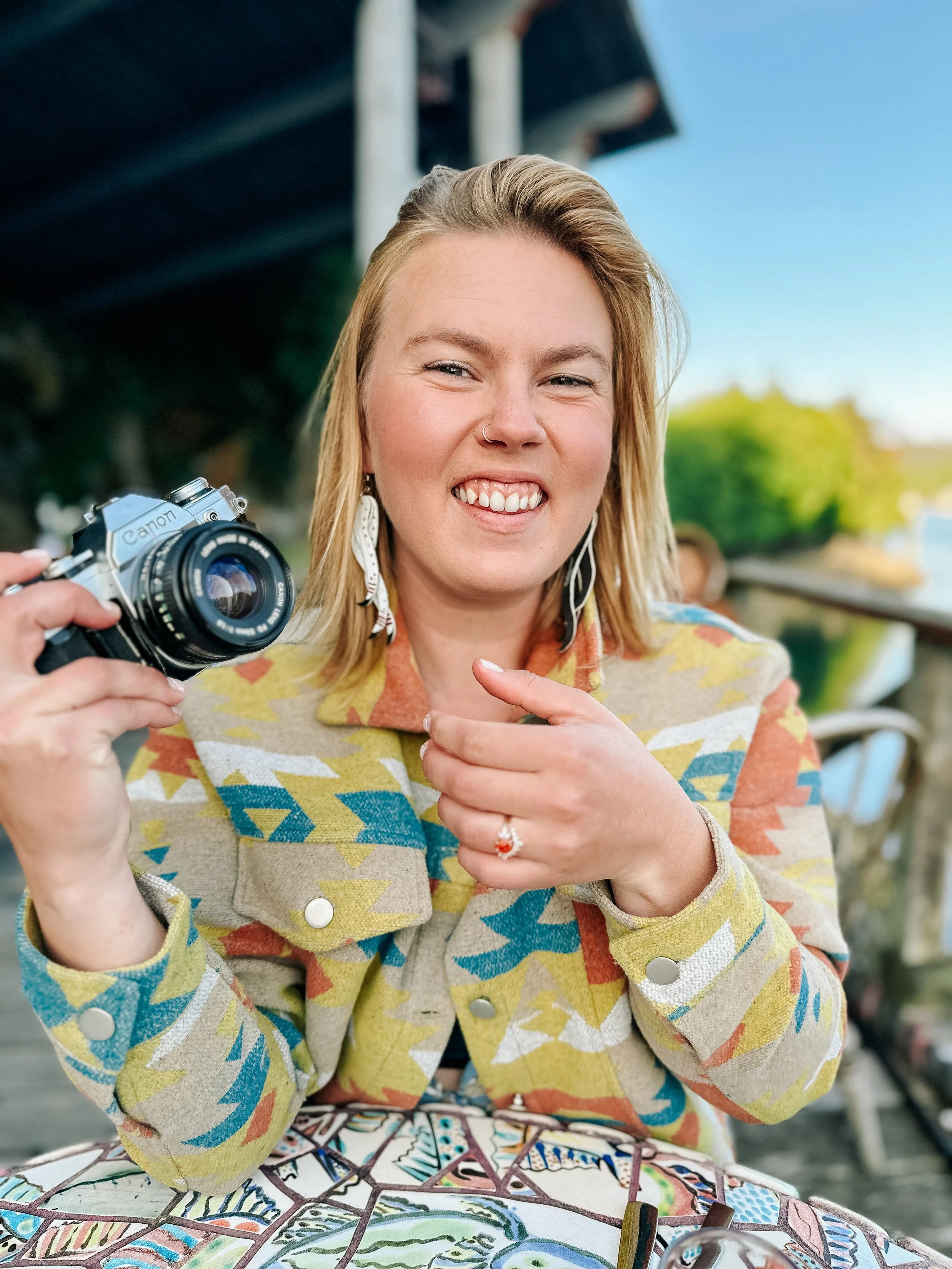 A woman smiling and pointing at a Canon camera she is holding, sitting at a colorful mosaic table outdoors with blurred background and blue sky.