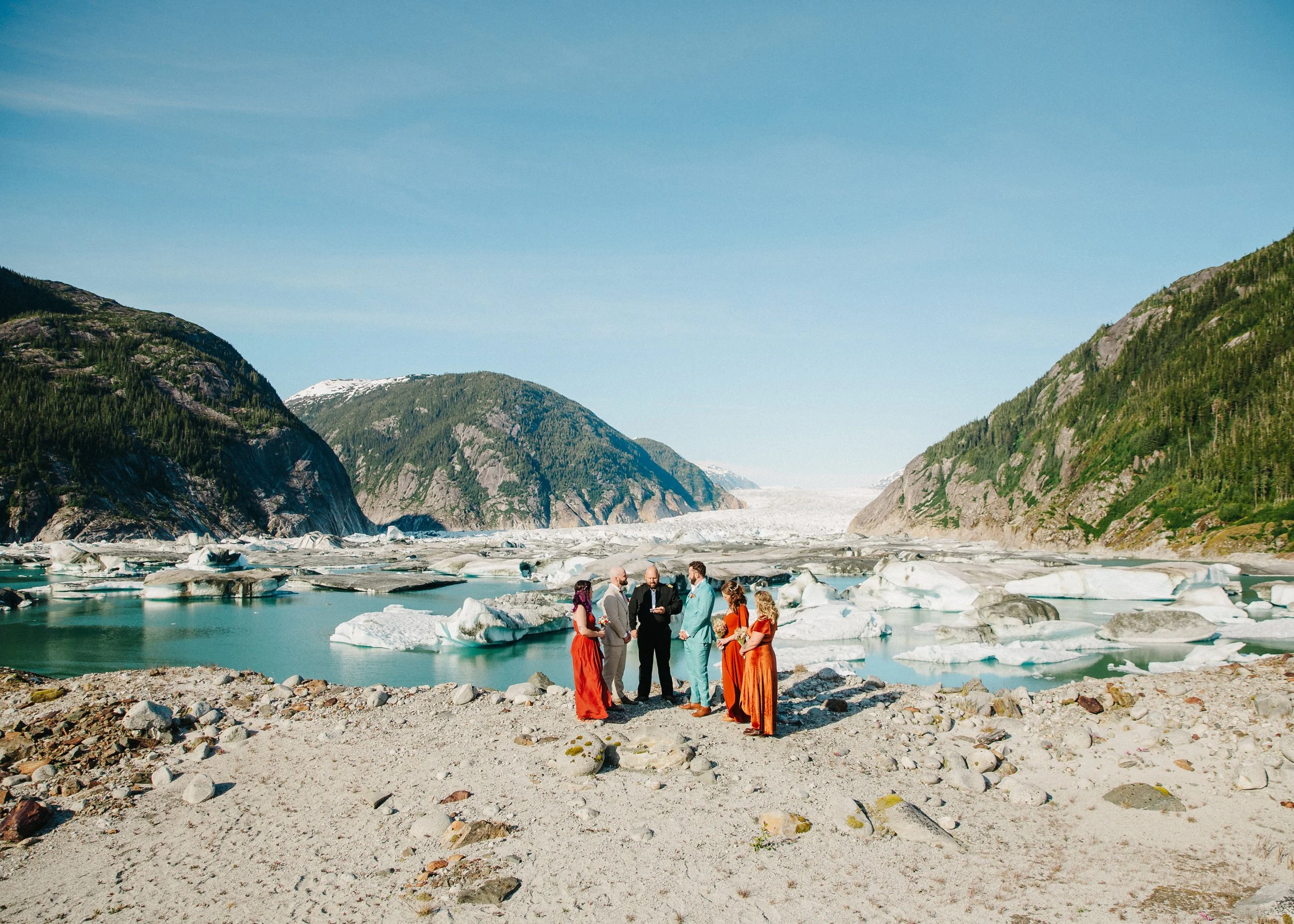 Group of people in wedding attire standing on rocky shore near glaciers and snow-capped mountains under clear blue sky.
