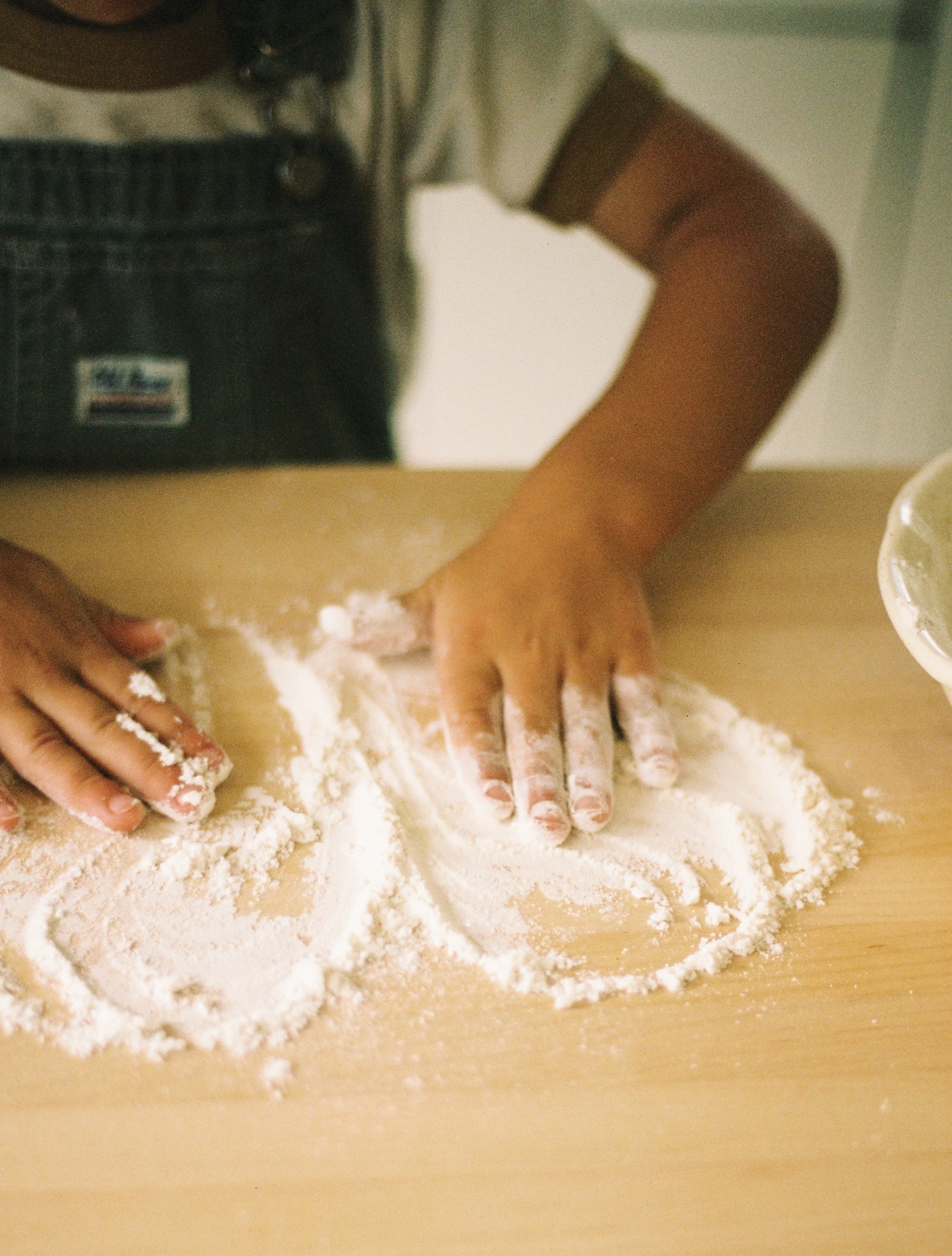 A closeup of a child's hands smoothing flour on a wooden table.