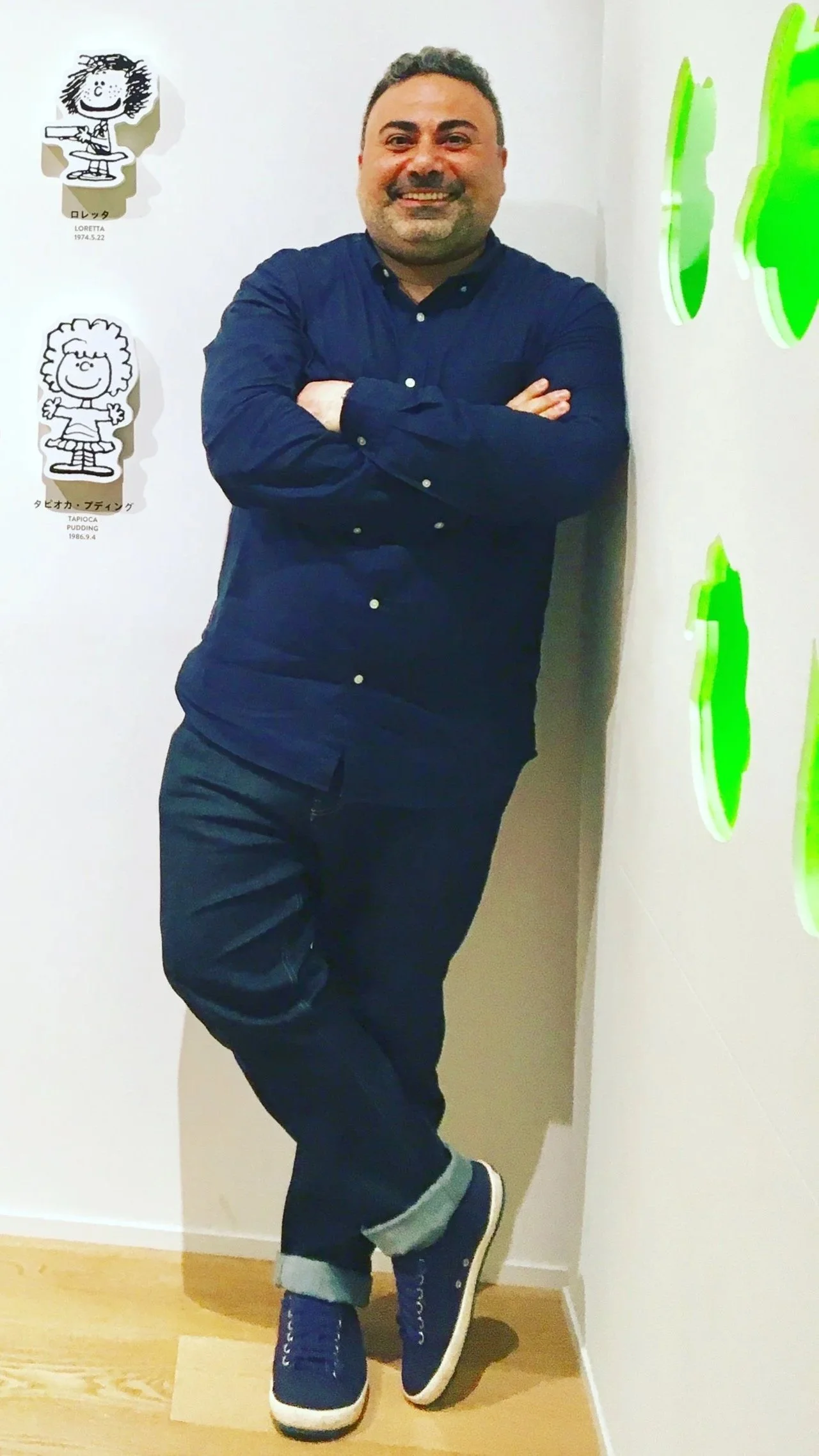 A person with short hair and a beard stands with arms crossed, smiling, wearing a dark long-sleeve shirt, dark pants, and blue sneakers, leaning against a white wall with cartoon character decals and bright green abstract wall art.