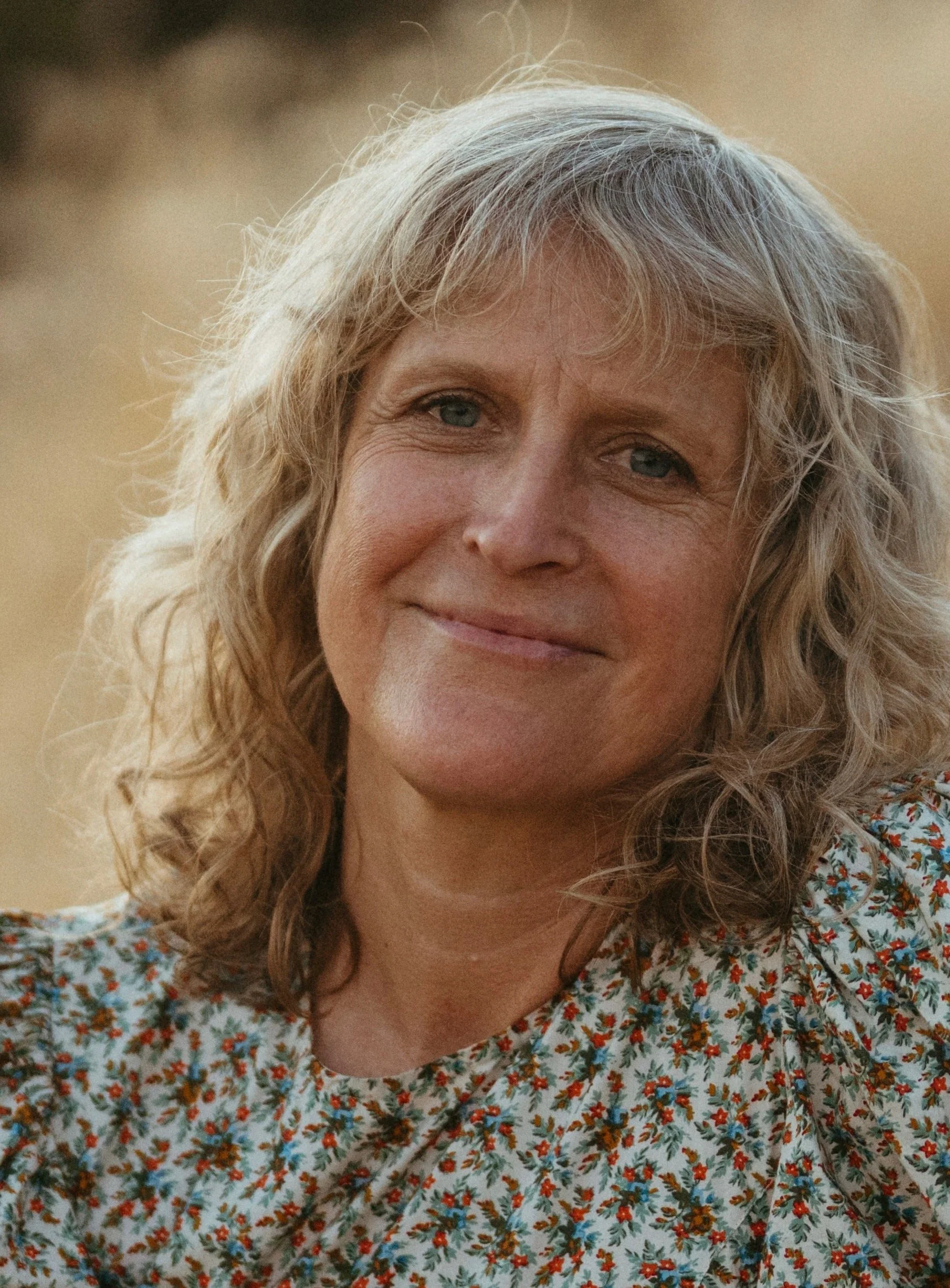 A woman with curly blonde hair and a floral dress sitting in tall dry grass during sunset with a background of rolling hills and trees.