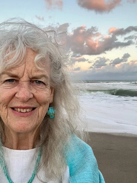 Older woman with long gray hair taking a selfie on a beach at sunset, with pink clouds and white capped waves in the background.
