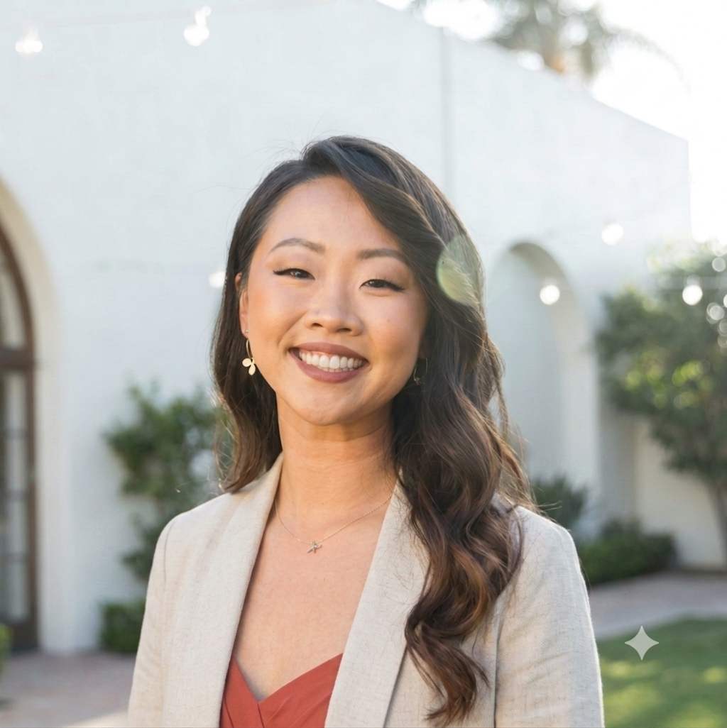 A woman with long wavy hair, smiling outdoors in front of a white building with arched windows and greenery.