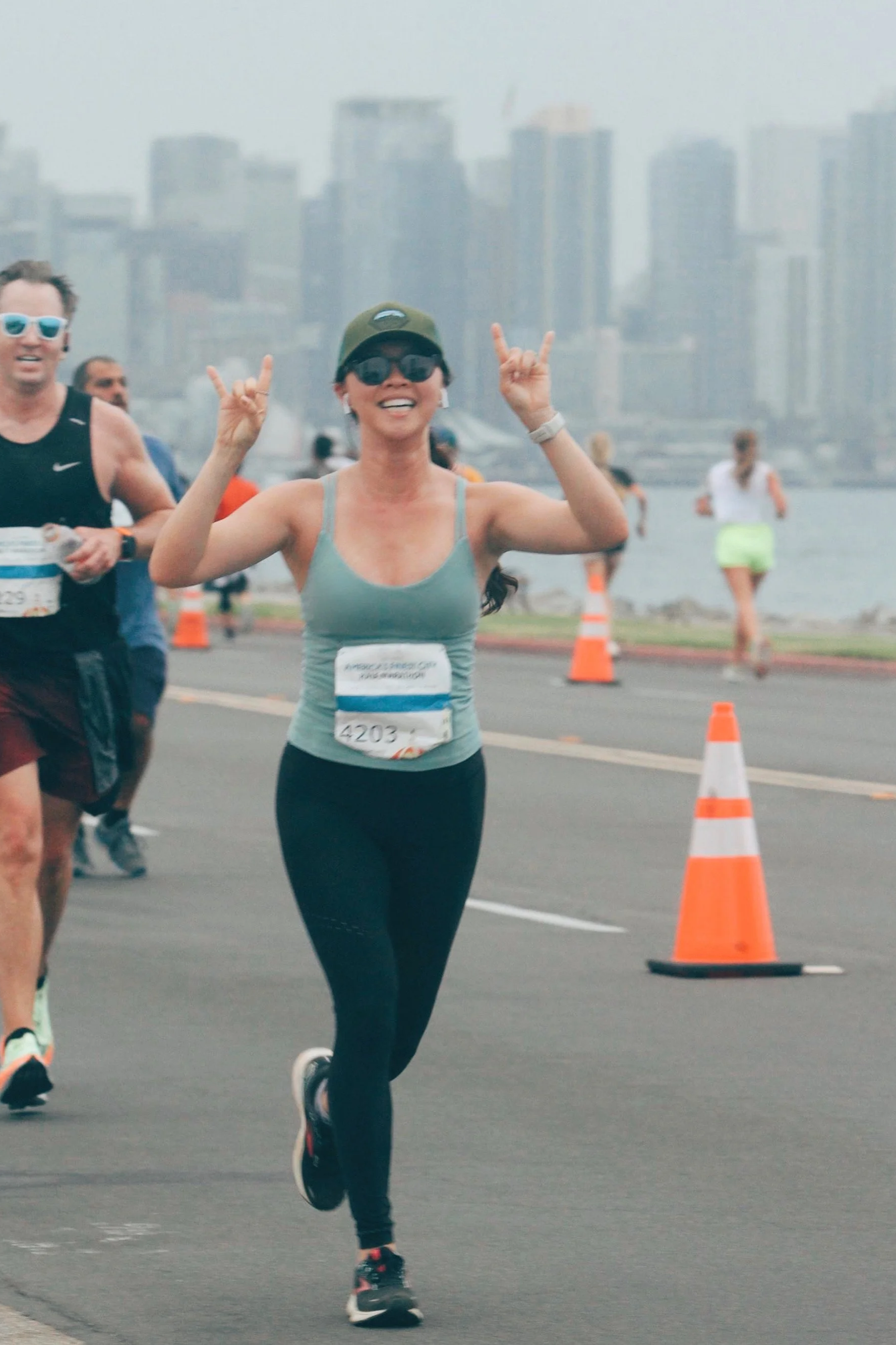 A woman running a marathon near a waterfront with a city skyline in the background. She is smiling, making a hand gesture, wearing sunglasses, a cap, a sleeveless top, and workout leggings.