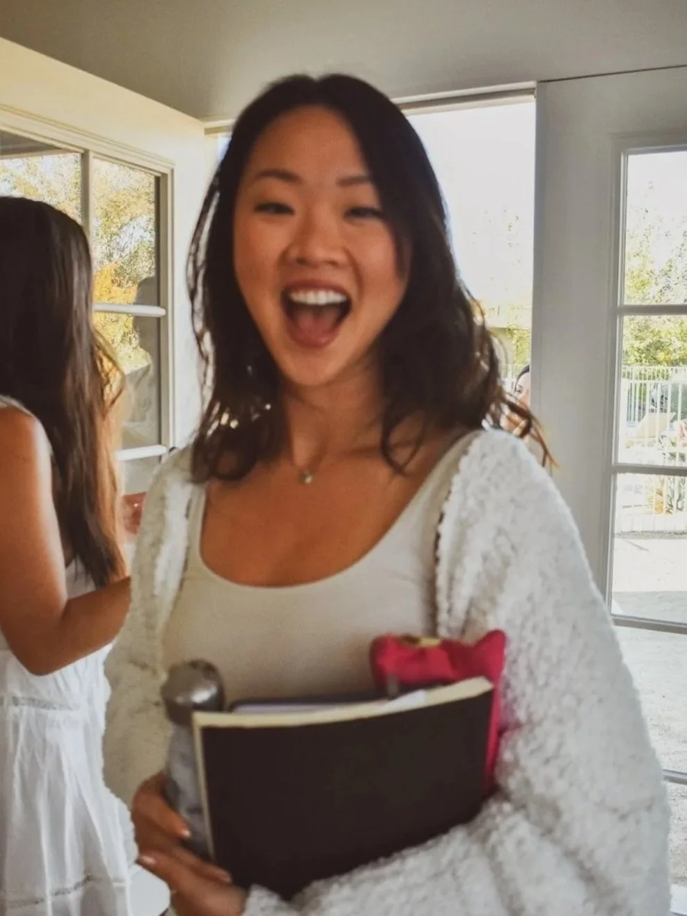 A young woman with black hair, smiling with her mouth open, holding a notebook and a pen, standing inside near a glass door with sunlight outside.