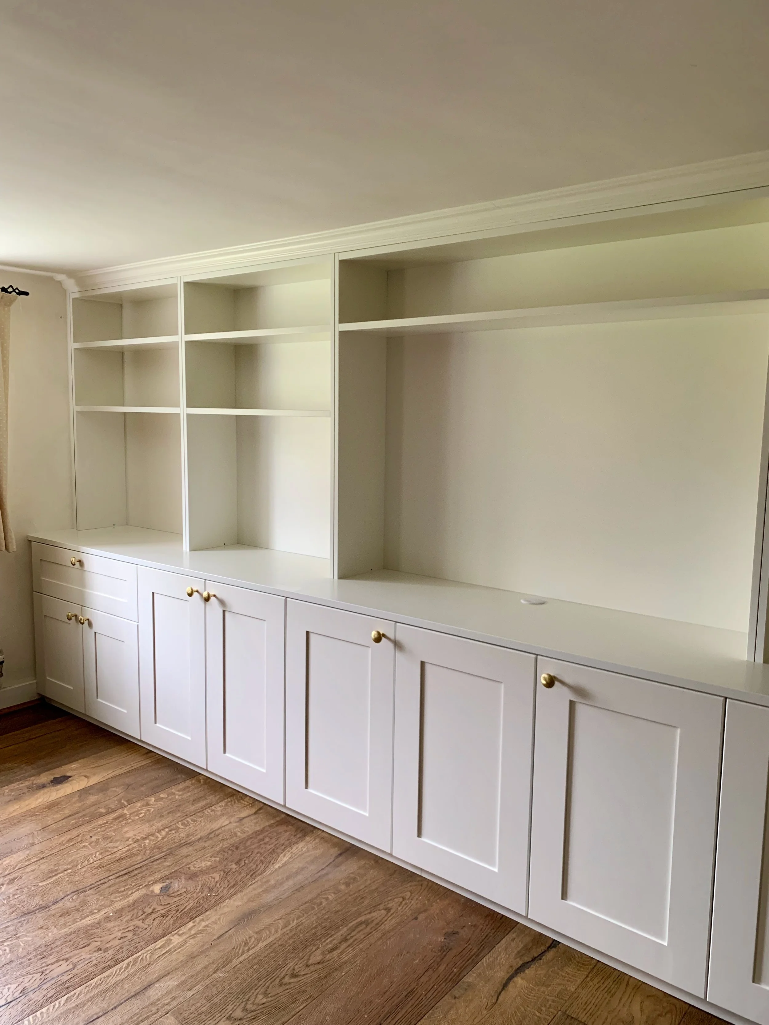 Empty white built-in bookshelf and cabinet unit with gold knobs in a room with wood flooring.