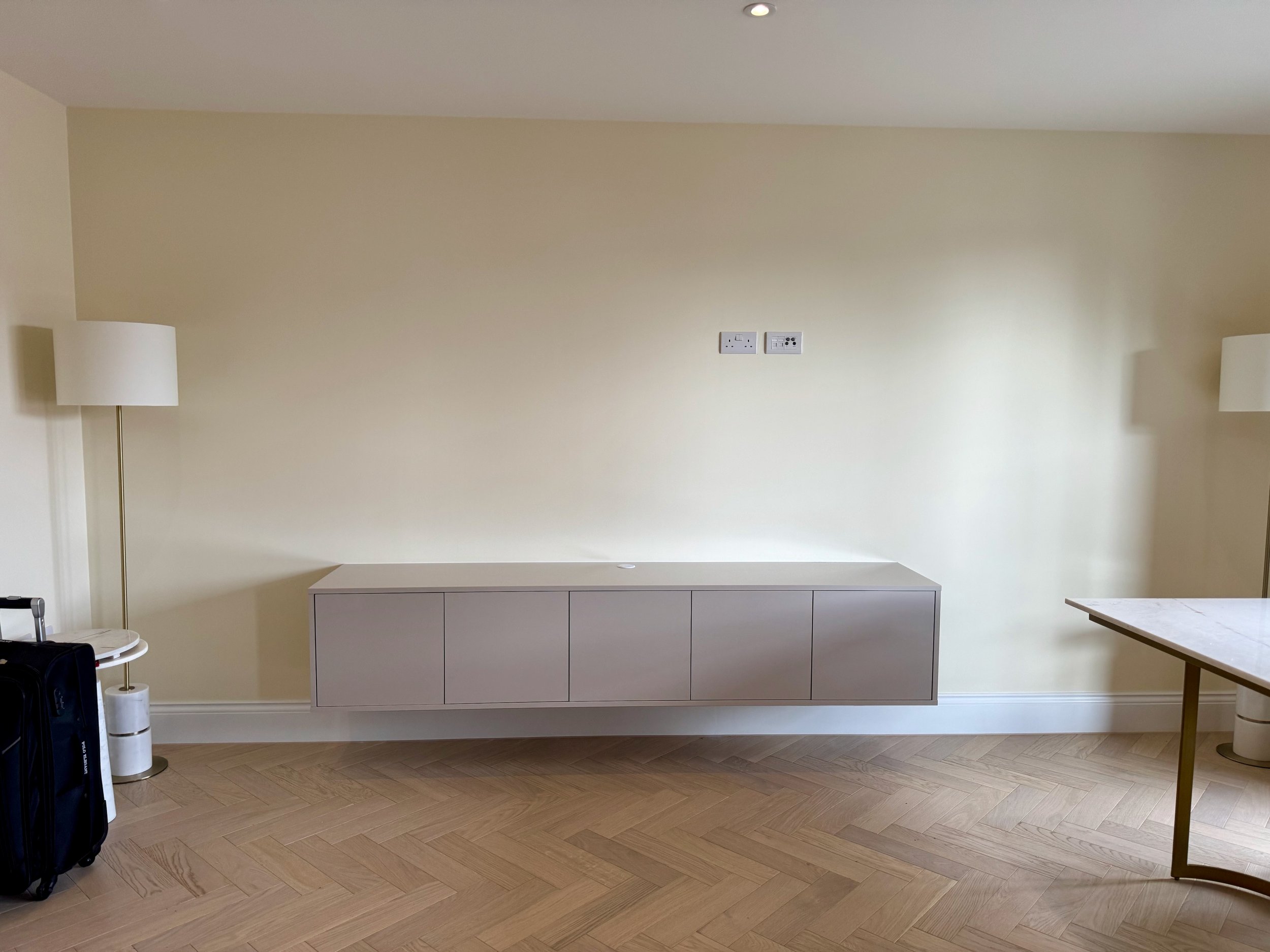 Minimalist hotel room with a floating gray cabinet on a beige wall, two tall floor lamps with white shades on each side, a small black suitcase, a white round table, and a marble-topped table with a gold frame.
