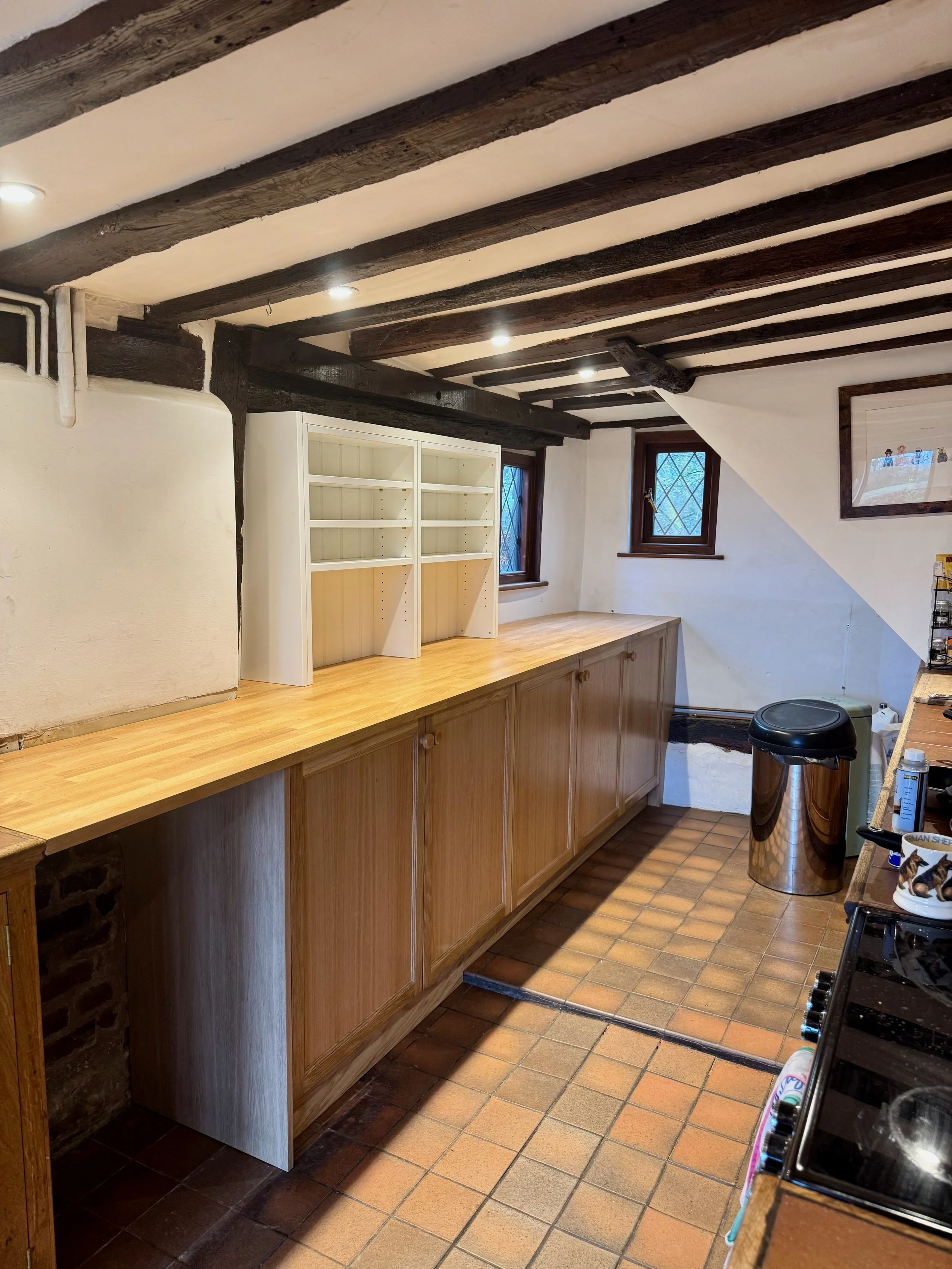 A cozy kitchen with exposed wooden beams on the ceiling, a long wooden countertop with cabinets, two small windows with diamond-shaped panes, and a metal trash can.
