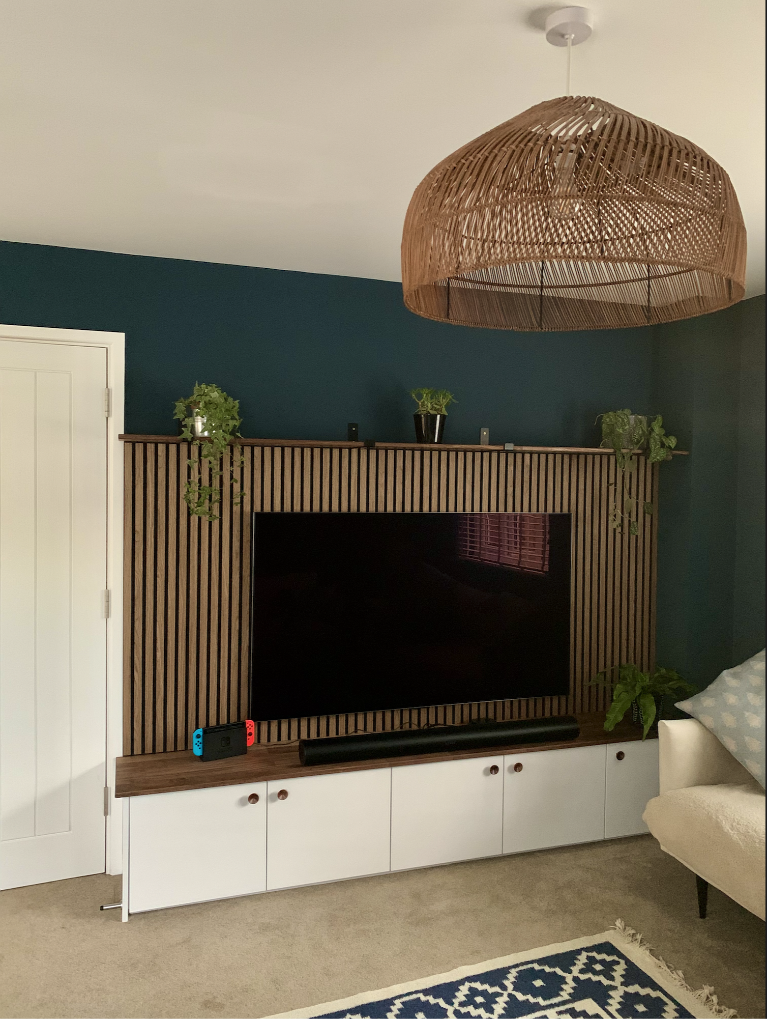 Living room with a wooden slatted panel behind a large flat-screen TV, green potted plants on a shelf above, a soundbar below, and an open door on the left. A couch with a patterned pillow is on the right, with a blue and white rug on beige carpet. A