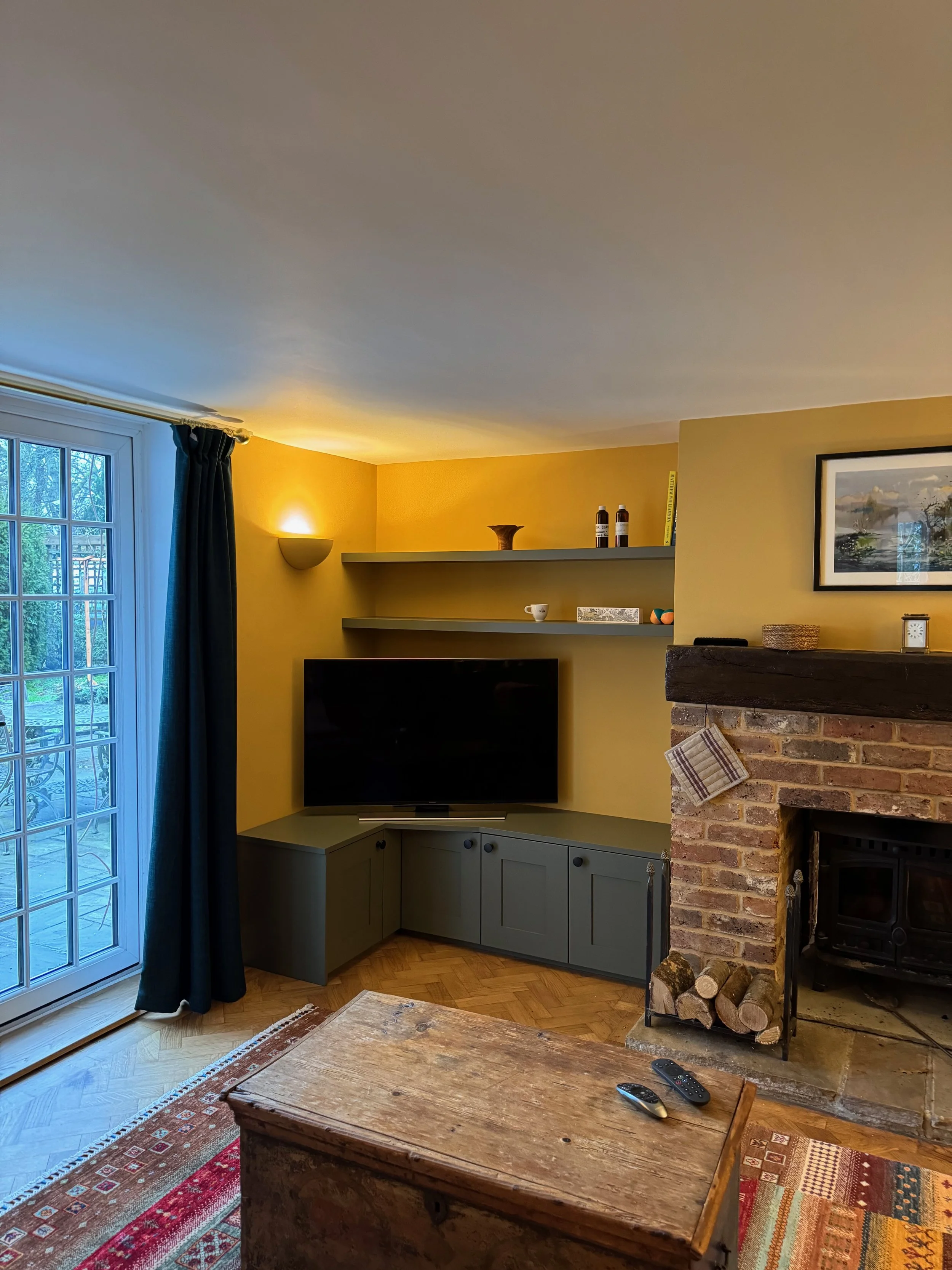 Living room with yellow accent wall, black flat-screen TV, green cabinetry below, floating shelves with decorative items, a brick fireplace with logs, a wooden coffee table, and a sliding glass door with dark curtains.