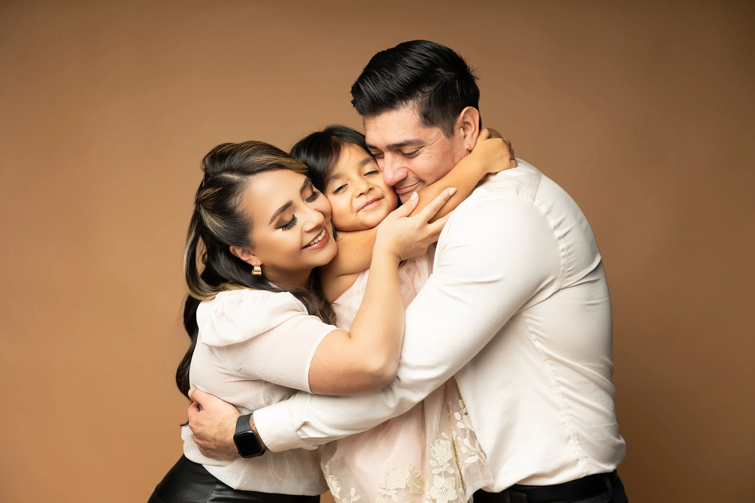 Family of three hugging each other and smiling, against a plain brown background.