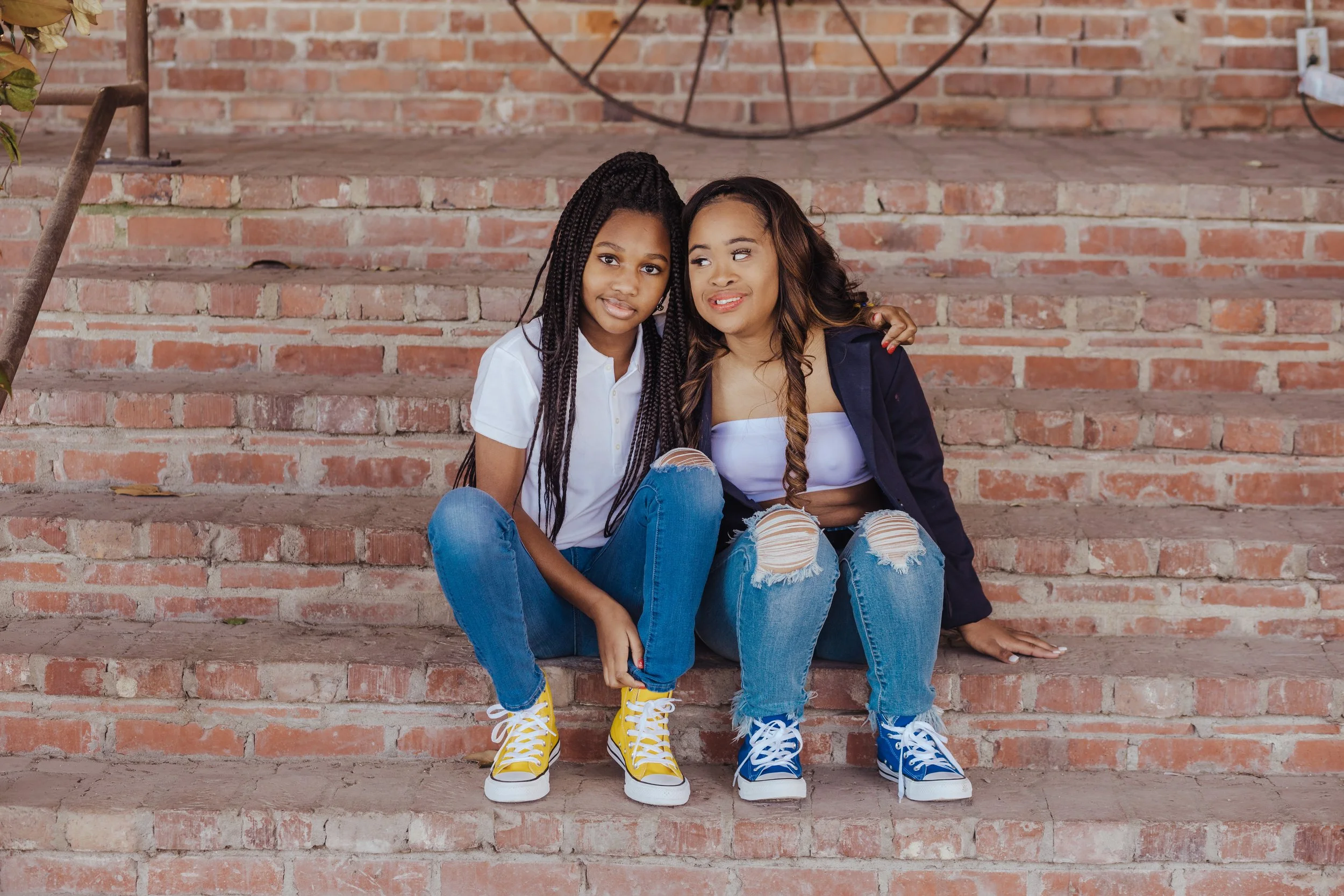 High school senior and her best friend sitting together on brick steps in Memphis, Tennessee during a senior portrait session by Hadonica’s Photography, wearing casual jeans and colorful sneakers, sharing a joyful and relaxed moment.