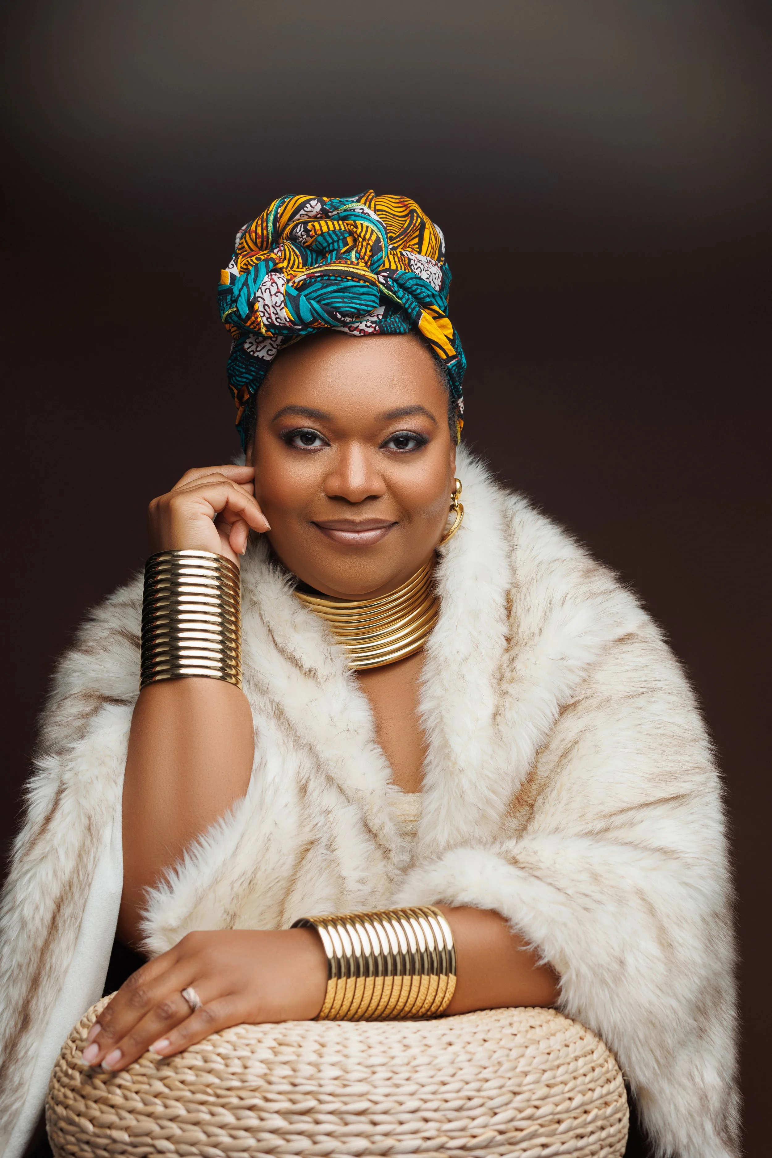Portrait of a woman wearing a colorful headwrap, gold jewelry, and a fur coat, sitting beside a woven basket against a dark background.