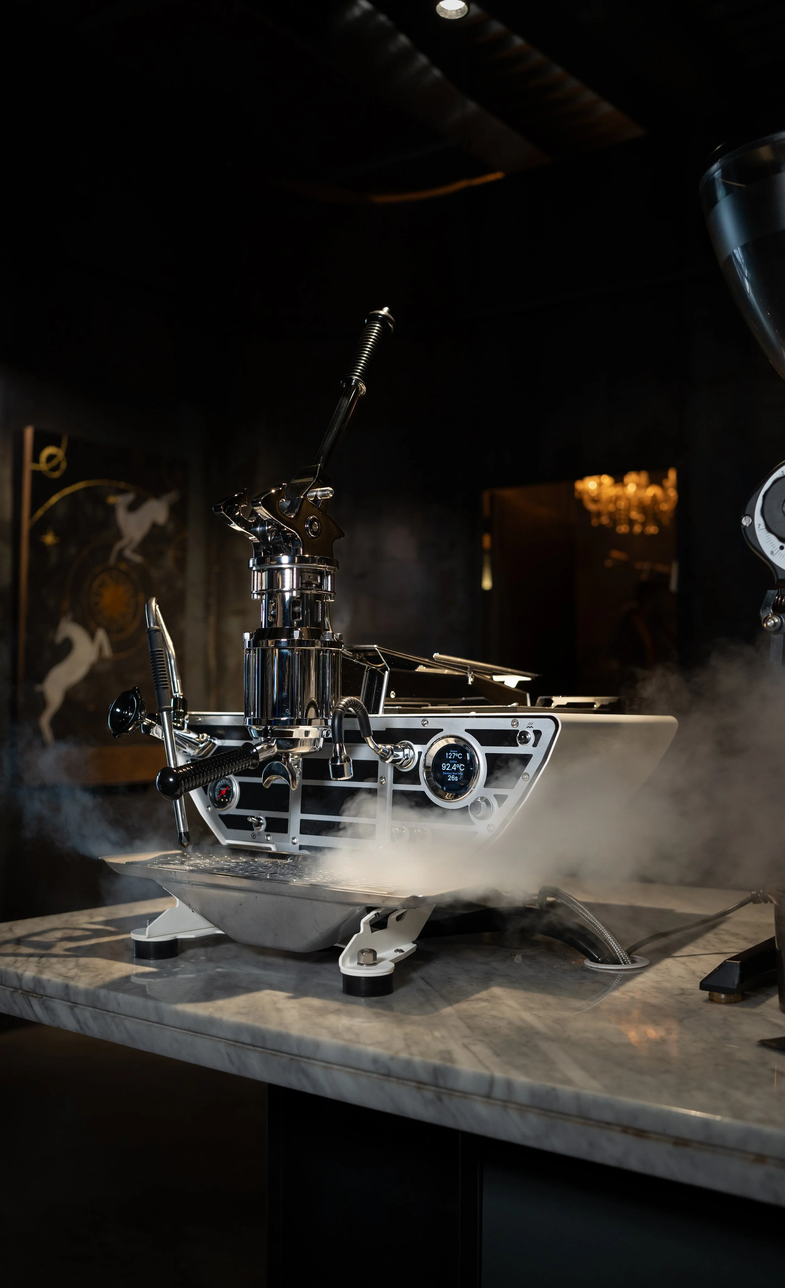A modern, high-tech coffee roaster with digital display on a marble countertop, emitting steam or smoke in a dark room.