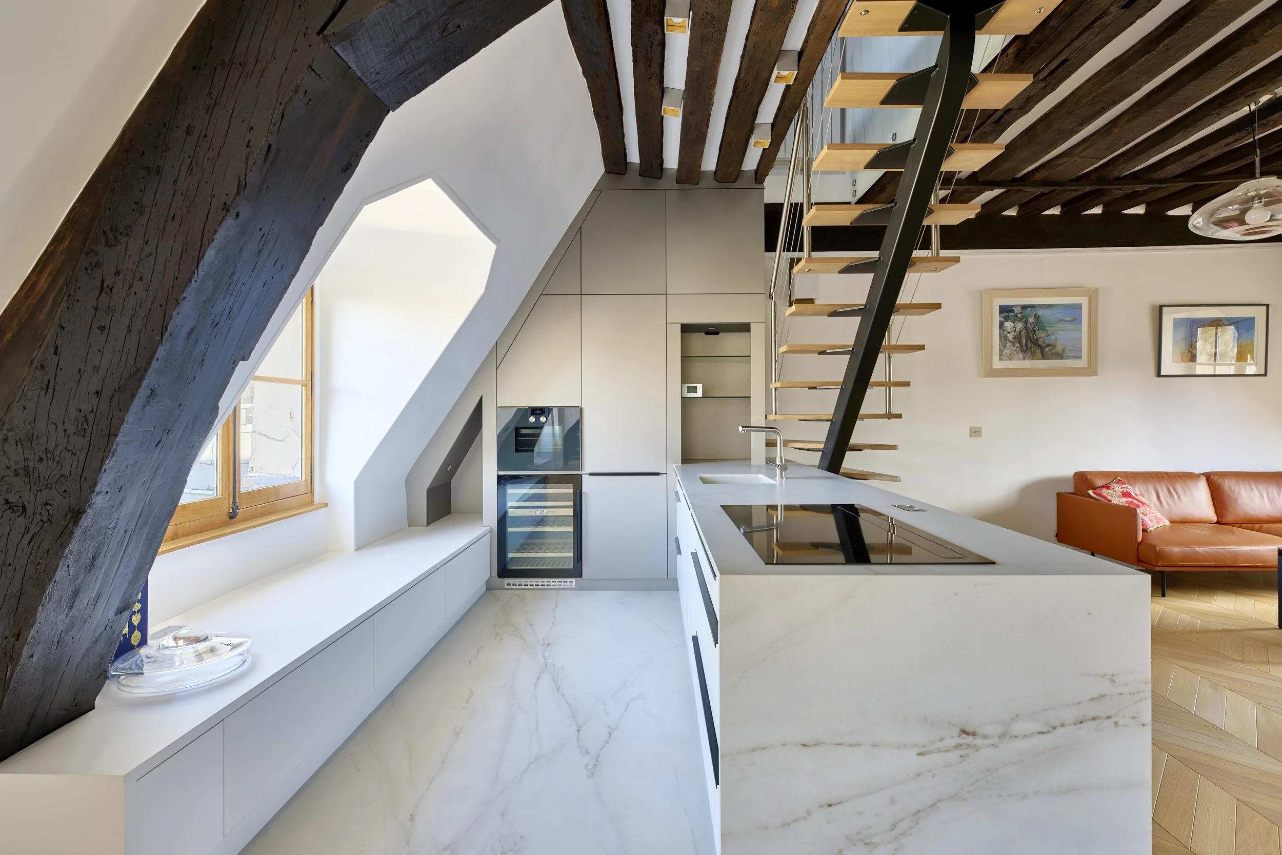 Modern kitchen with white marble countertops, a built-in oven, and a small wine cooler. Large window with wooden trim, exposed dark wood beams, and a staircase with wooden steps and black metal supports leading to an upper floor. Adjacent living area