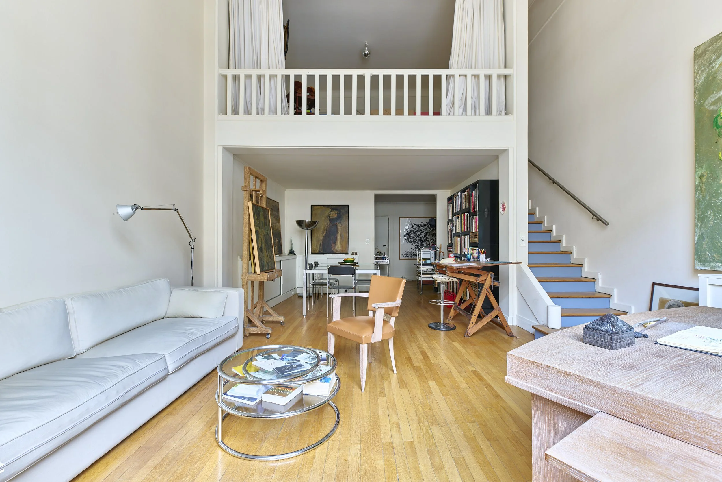 Interior of a spacious living room with a white couch, glass coffee table, wooden floors, and a loft area with white curtains. There are chairs, bookshelves, and artwork on the walls.