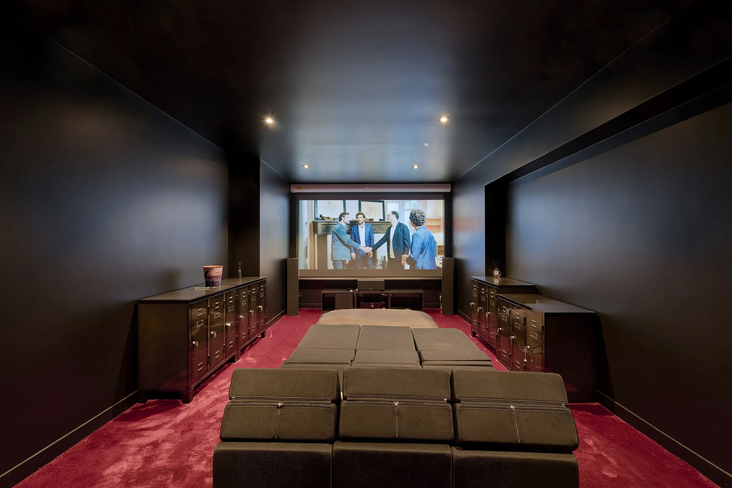 Home theater room with black walls, red carpet, a large screen showing a business handshake, and black cabinets along the sides.