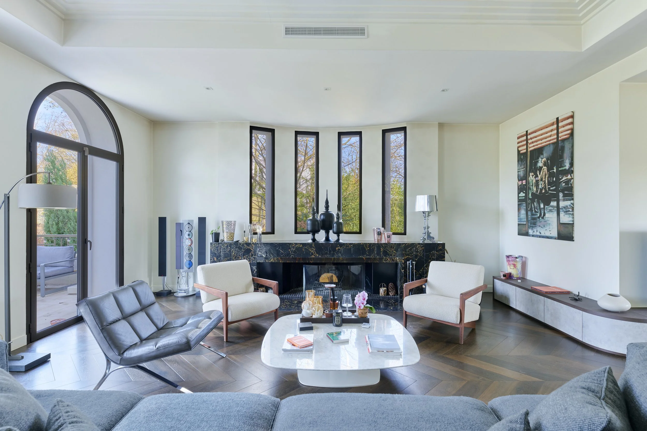 Modern living room with a fireplace, two beige armchairs, a black leather lounge chair, a white coffee table with books and decorative items, artwork on the wall, and large windows showing trees outside.