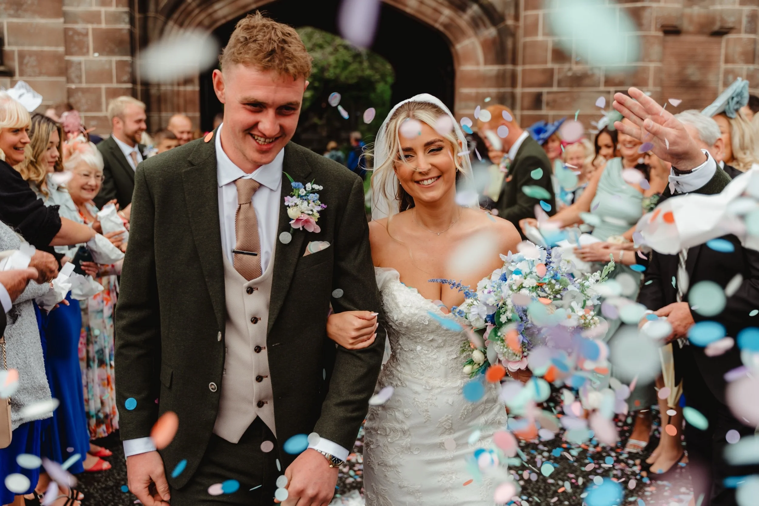 A bride and groom walking arm in arm through a shower of colorful confetti, surrounded by smiling guests outside a church.