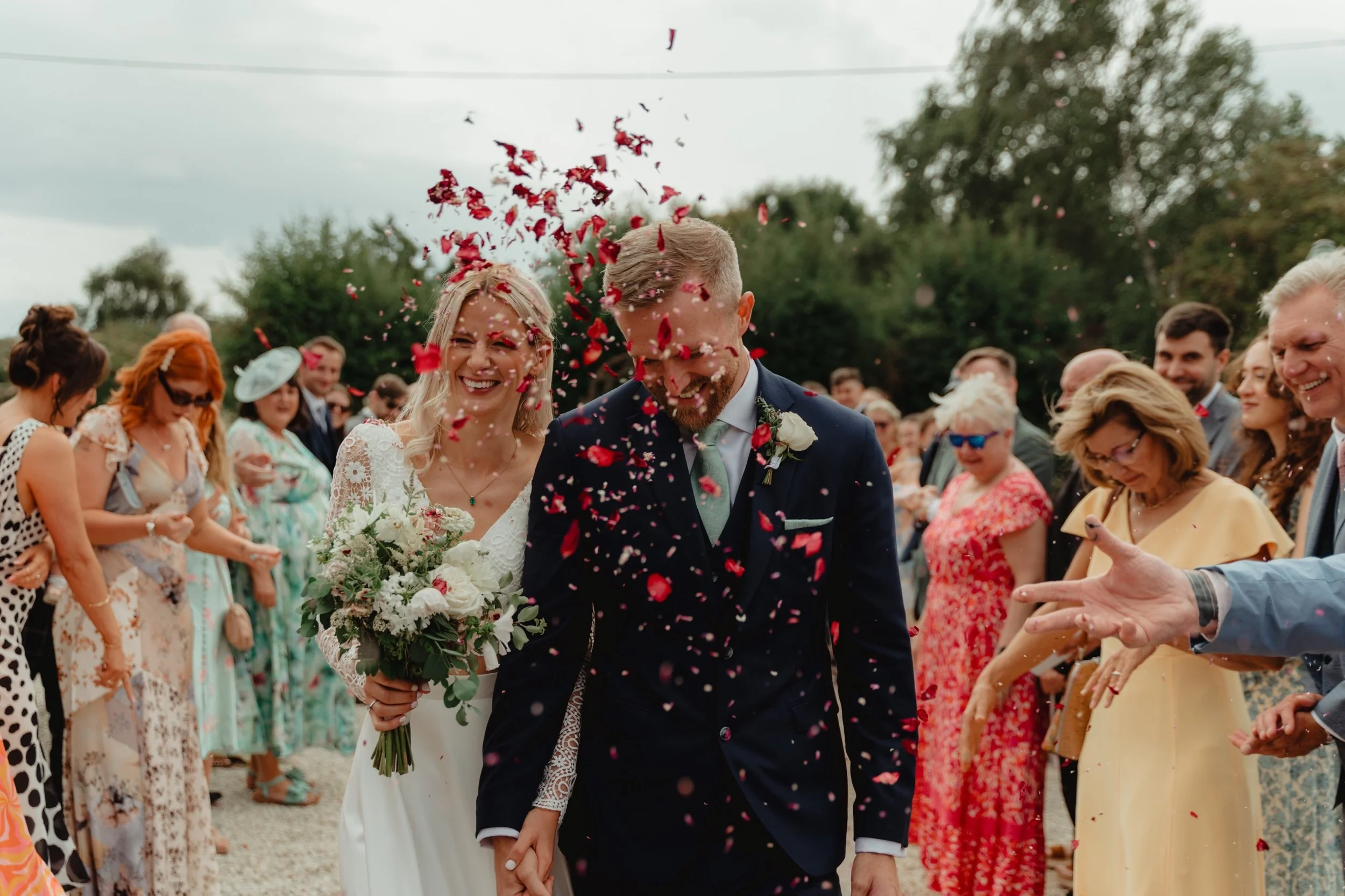 A bride and groom smiling as they walk through confetti during their outdoor wedding celebration, surrounded by guests.