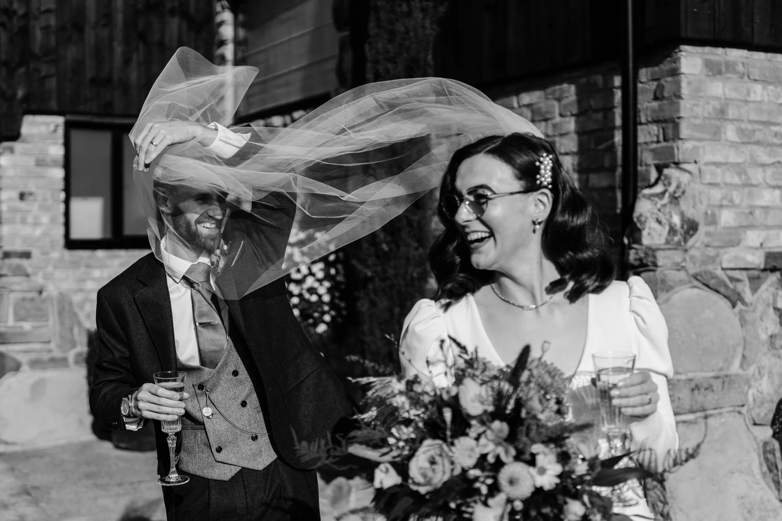 A happy bride and groom sharing a joyful moment outdoors, with the groom lifting a veil over the bride her holding a bouquet, both smiling and celebrating.