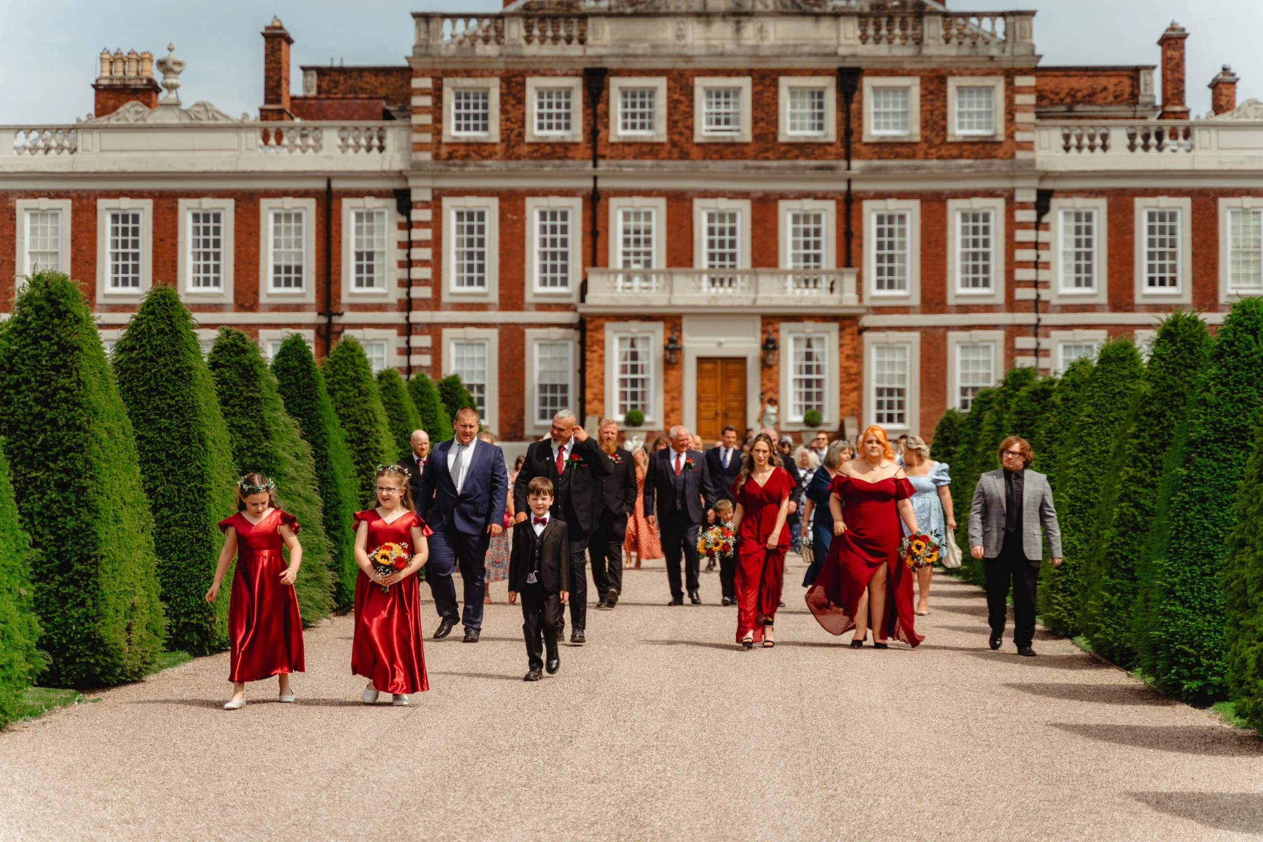 A wedding party walking in front of a large red-brick mansion, with green hedges lining the gravel pathway. The group includes children in red dresses, adults in formal attire, and some holding bouquets of flowers.